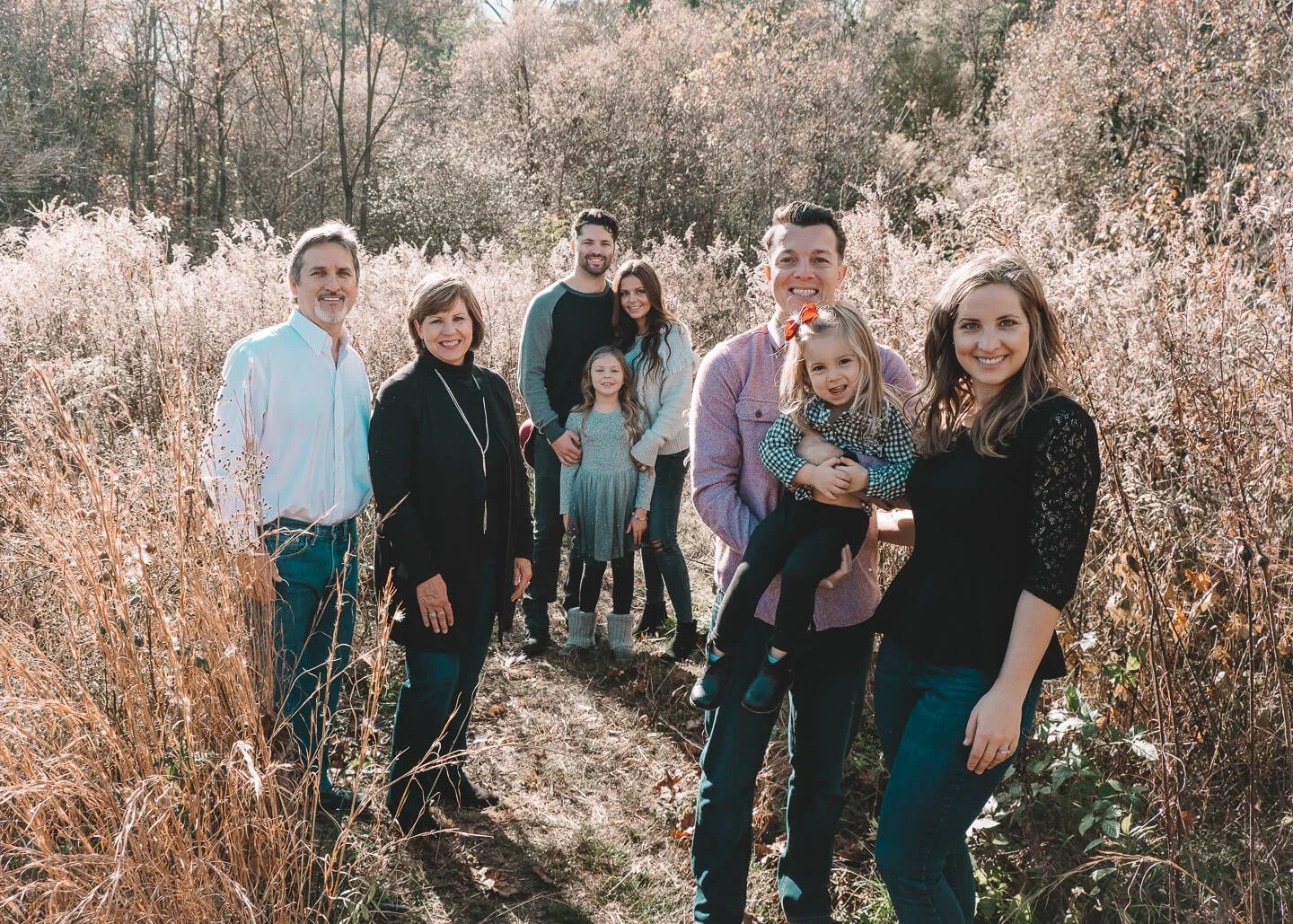 A family of eight standing together in a field during fall, smiling at the camera.