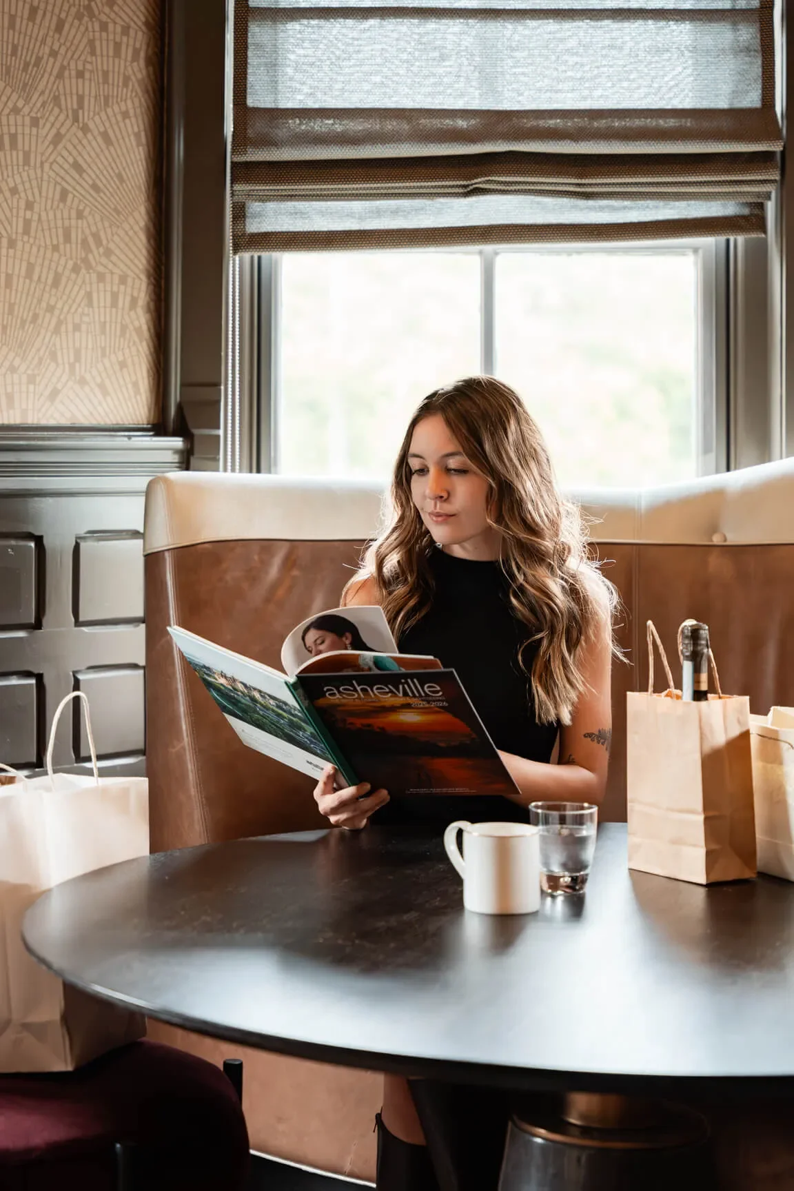 A young woman with long wavy hair sitting at a booth table in a restaurant, reading a magazine titled 'asheville'. There are paper bags, a coffee mug, and a glass of water on the table, with a window behind her letting in natural light.