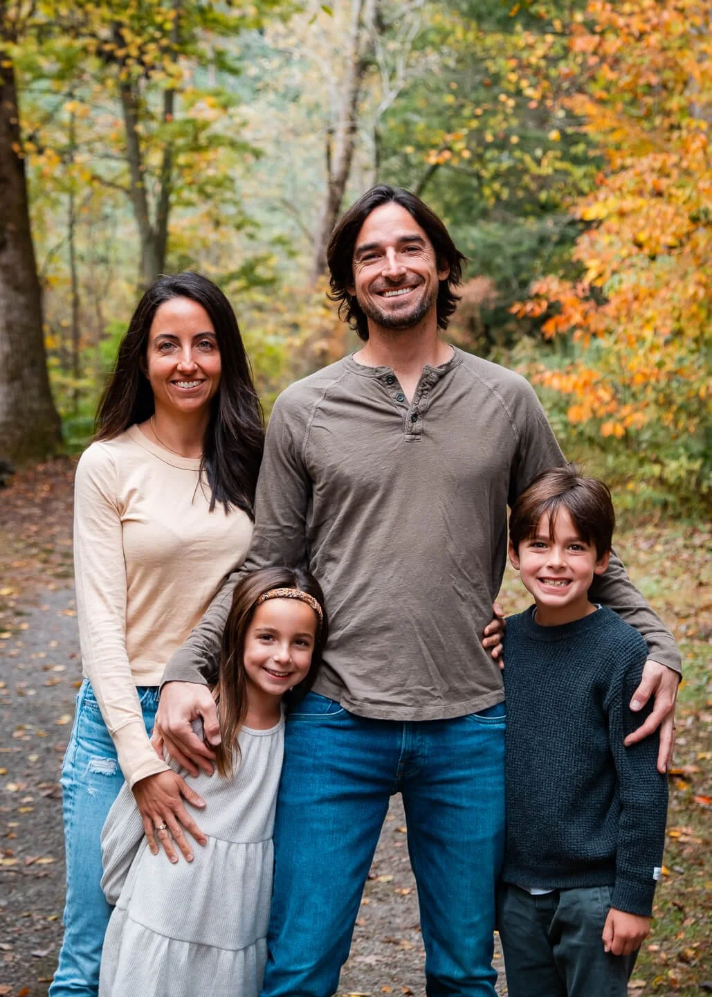 Family of four standing together outdoors in an autumn forest, smiling at the camera. The father and mother are behind their daughter and son, who are in front, with colorful fall leaves in the background.