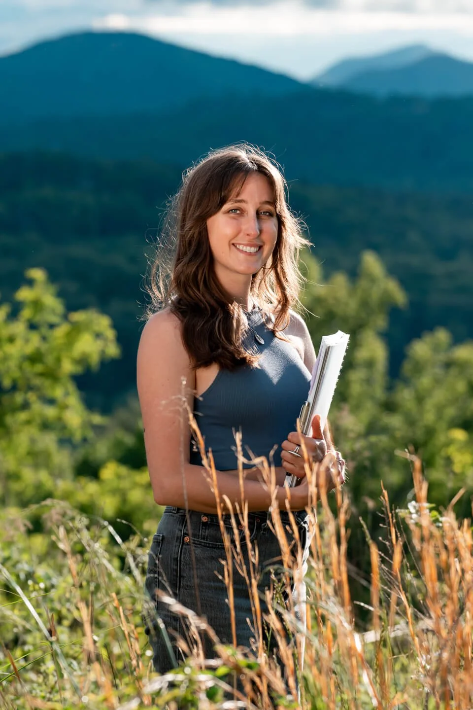 A young woman with long brown hair smiling while holding notebooks in a field with tall grass, set against a backdrop of green mountains under a partly cloudy sky.