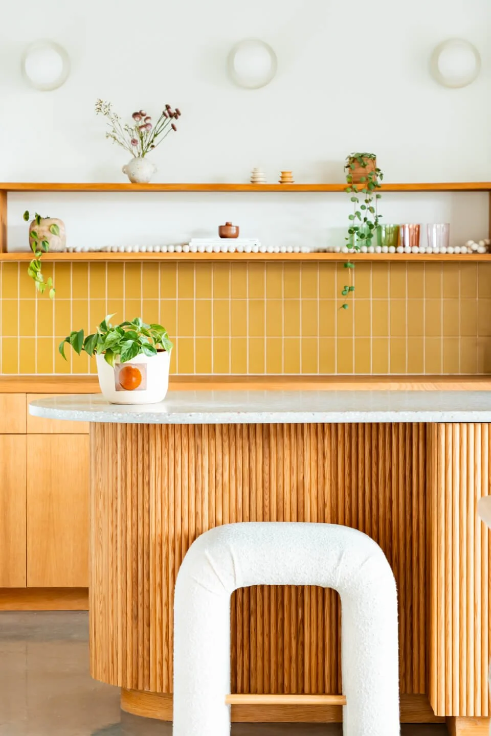 Interior of a modern kitchen with a white countertop, wooden cabinetry, yellow tiled backsplash, and decorative items including plants and ceramics on open wooden shelves.