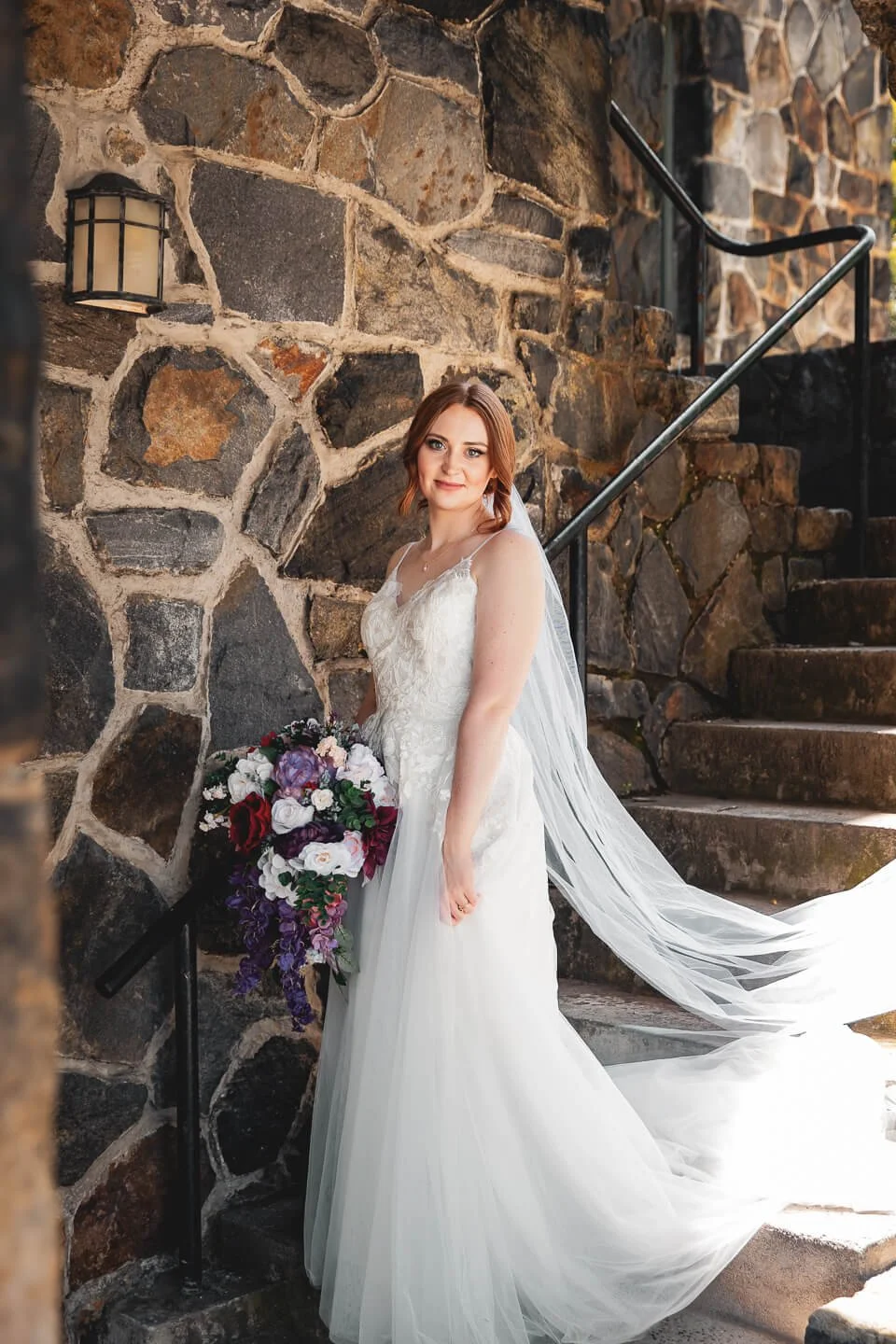 A bride in a white lace wedding dress with a long veil, holding a large bouquet of purple, white, and red flowers, standing on stone stairs next to a stone wall, smiling at the camera.