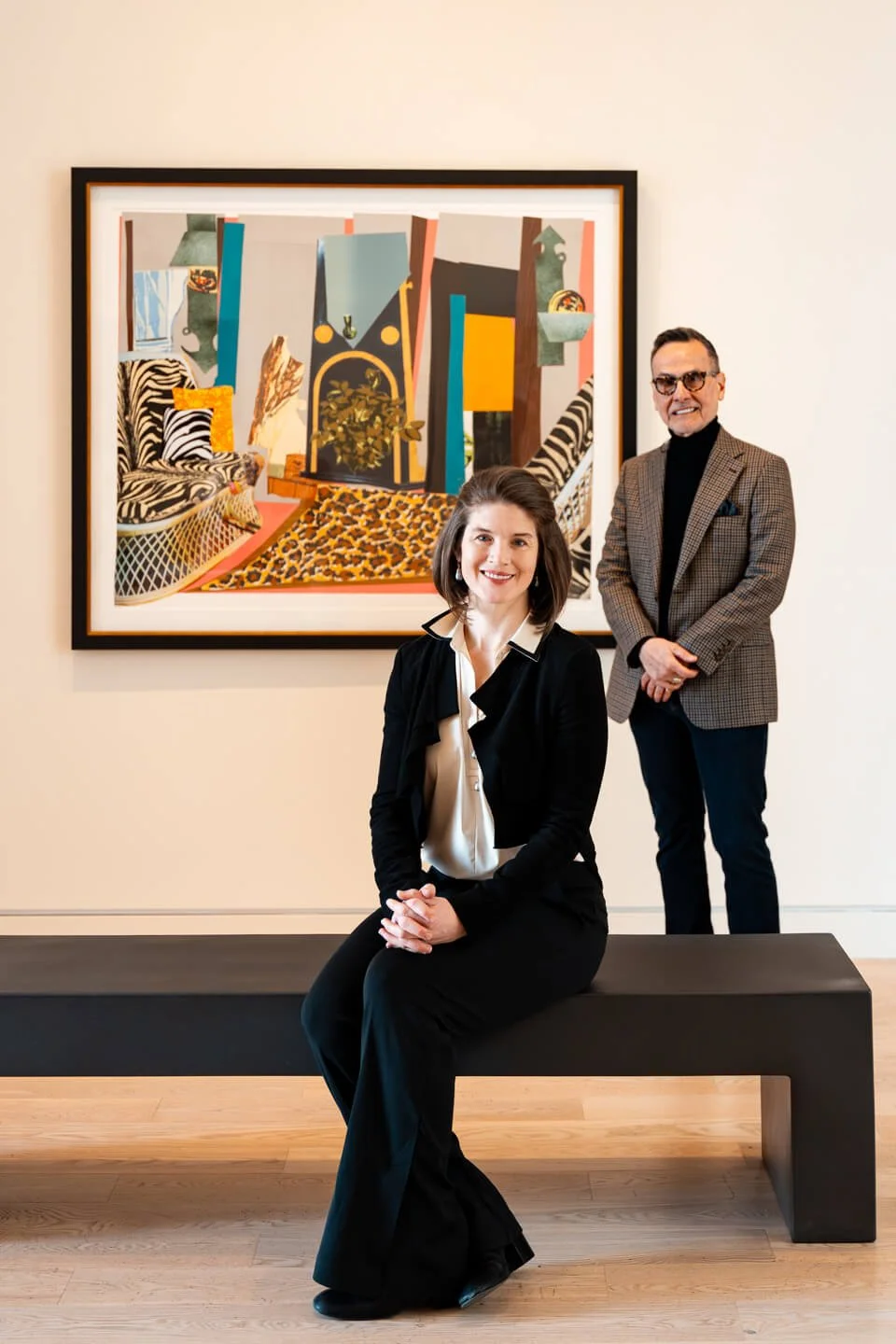 A woman is sitting on a black bench, smiling, with a man standing behind her to the right, both dressed professionally, in an art gallery with an abstract painting on the wall behind them.