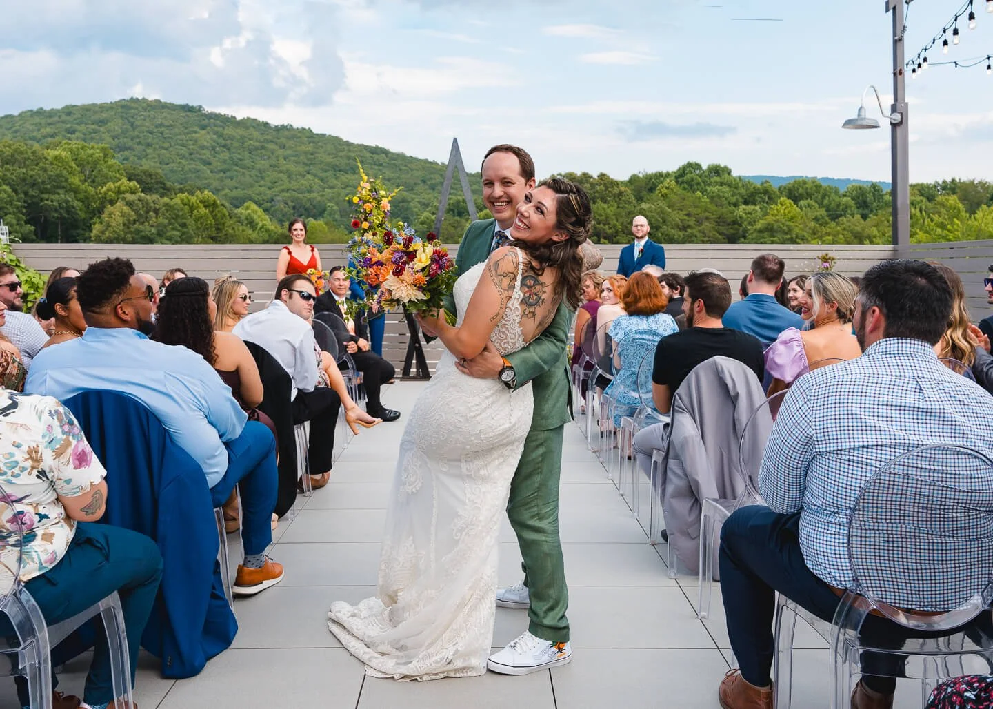 A newly married couple hugging during an outdoor wedding ceremony, with guests seated on either side and scenic mountains in the background.