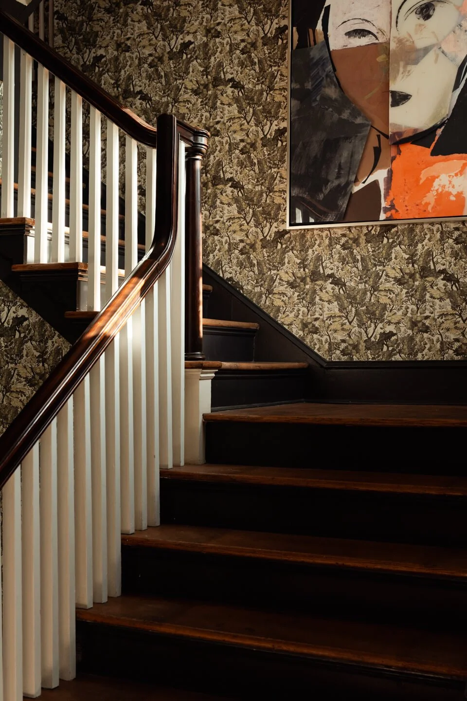 Interior view of a staircase with dark wooden steps, white spindles, and a dark brown handrail, against wallpaper with a tree pattern and a large colorful portrait painting on the wall.