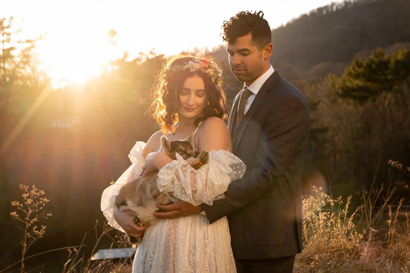 A bride and groom holding a small goat during sunset in a rural outdoor setting with mountains and trees in the background.