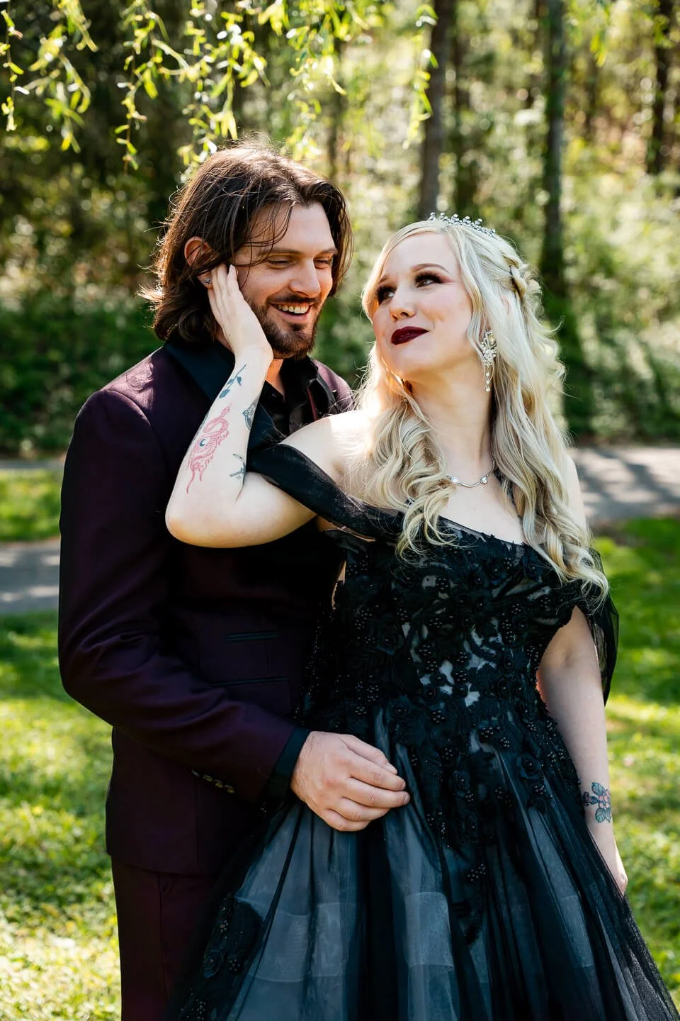 A couple standing close together outdoors, smiling; the woman has long blonde hair, dark lipstick, and tattoos, wearing a black dress with lace details. The man has long hair, a beard, and is dressed in a dark suit. They are surrounded by green trees