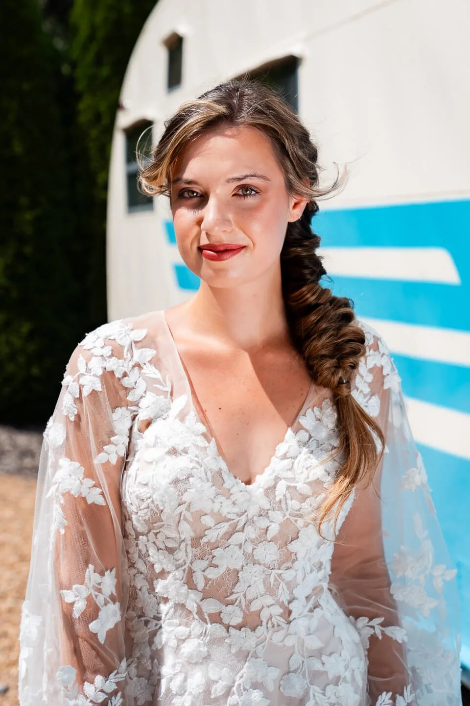 Young woman with light brown hair in a loose braid, wearing a sheer white lace floral dress, standing outdoors in bright sunlight with a modern building and blue and white striped surface in the background.
