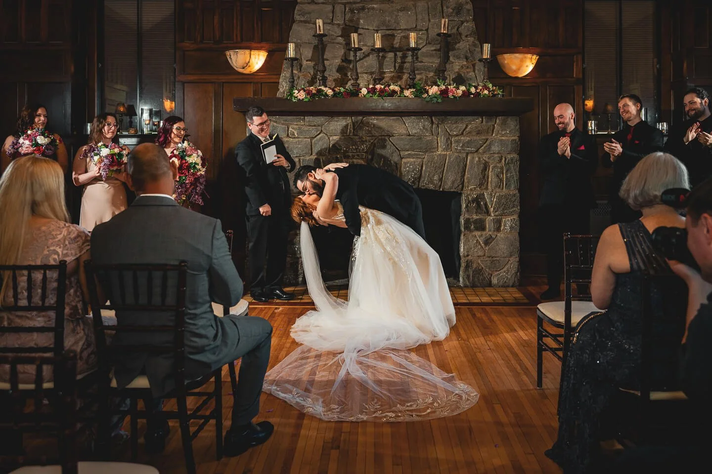 Bride and groom sharing a kiss during their wedding ceremony in front of a stone fireplace, surrounded by guests and wedding party in formal attire.