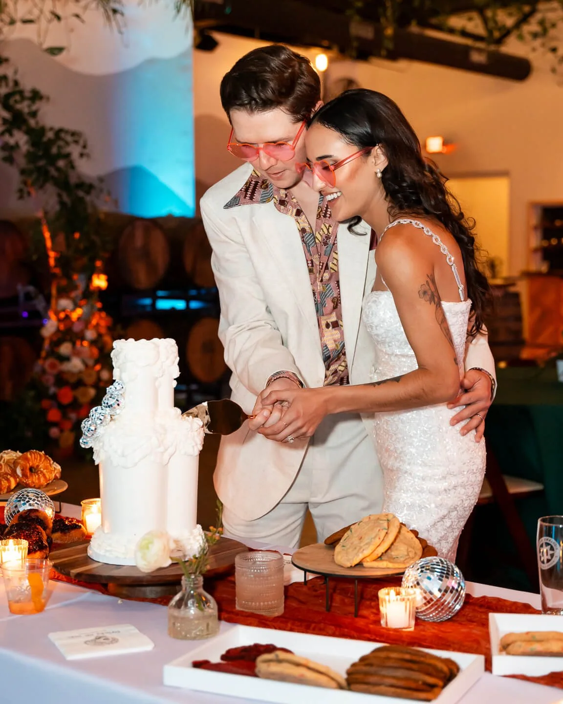A bride and groom cutting a wedding cake together at their reception, smiling and holding a cake knife, surrounded by desserts and candles.