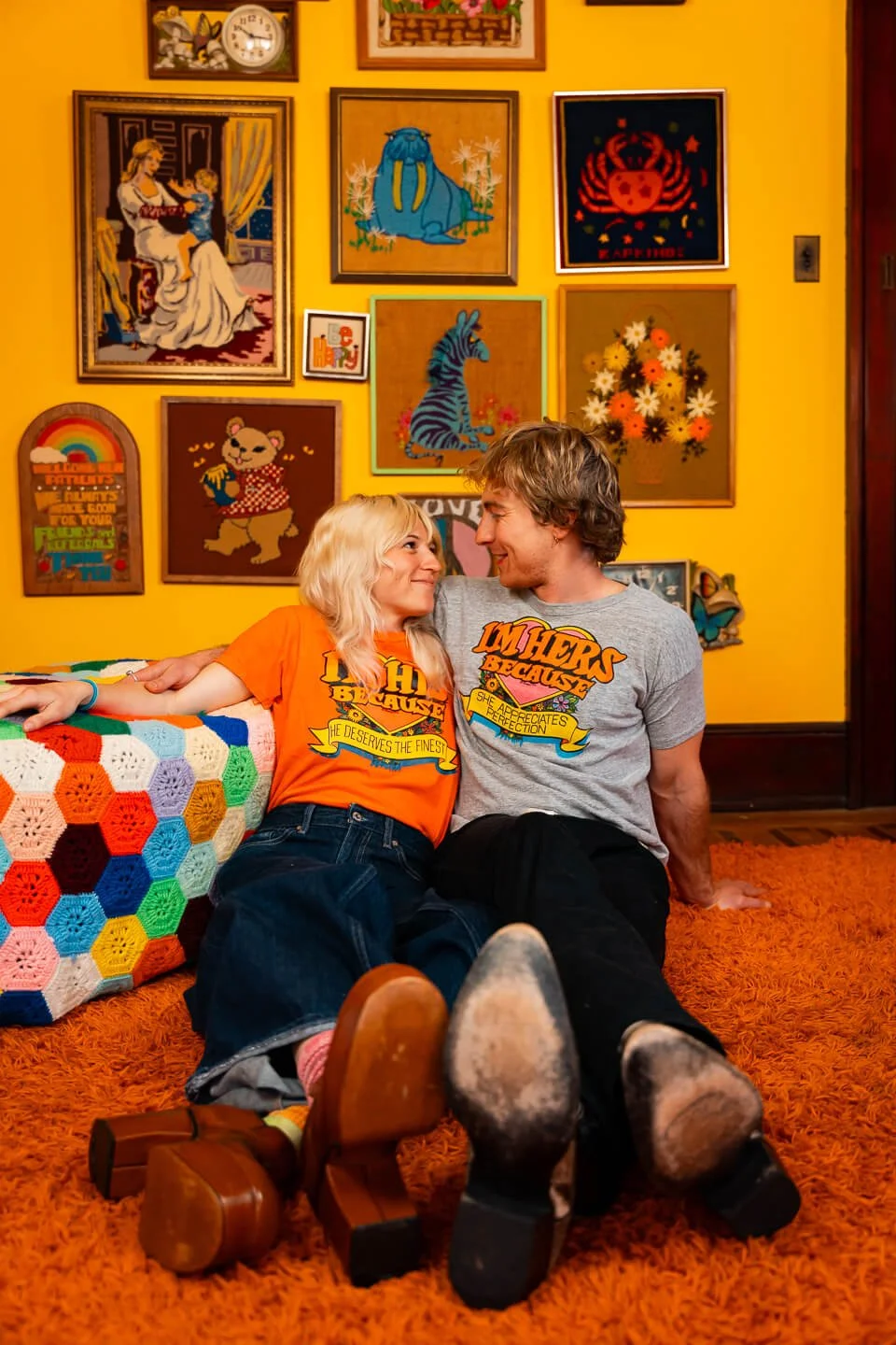 A young woman and a young man sit on an orange shag carpet, leaning against a multicolored crocheted blanket. They are smiling at each other, wearing matching T-shirts with colorful text and designs. The background features a yellow wall decorated wi