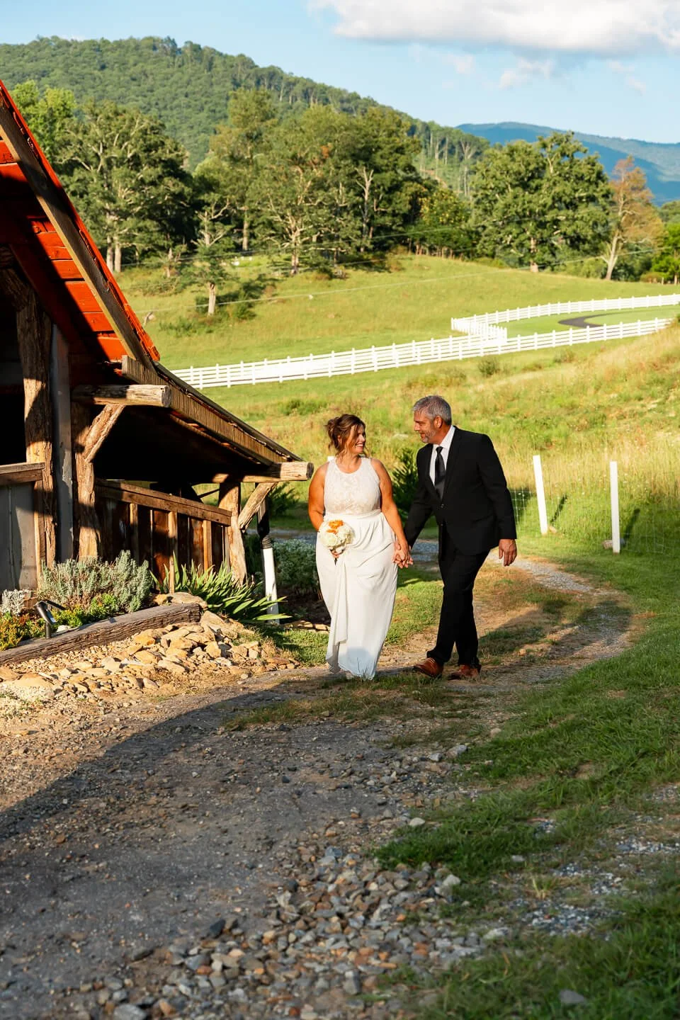 A bride and groom walk hand in hand outdoors on a gravel path, smiling at each other. The bride wears a white wedding dress and holds a bouquet, while the groom is dressed in a black suit and tie. The background features rolling green hills, trees, a