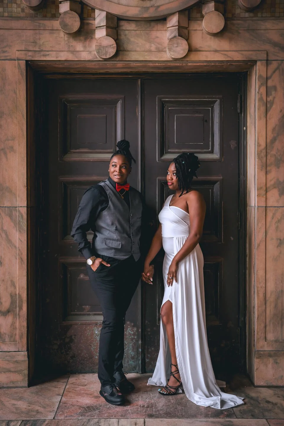 Two women holding hands in front of a large wooden door, one in a tuxedo with a red bow tie and the other in an elegant white dress with a slit.
