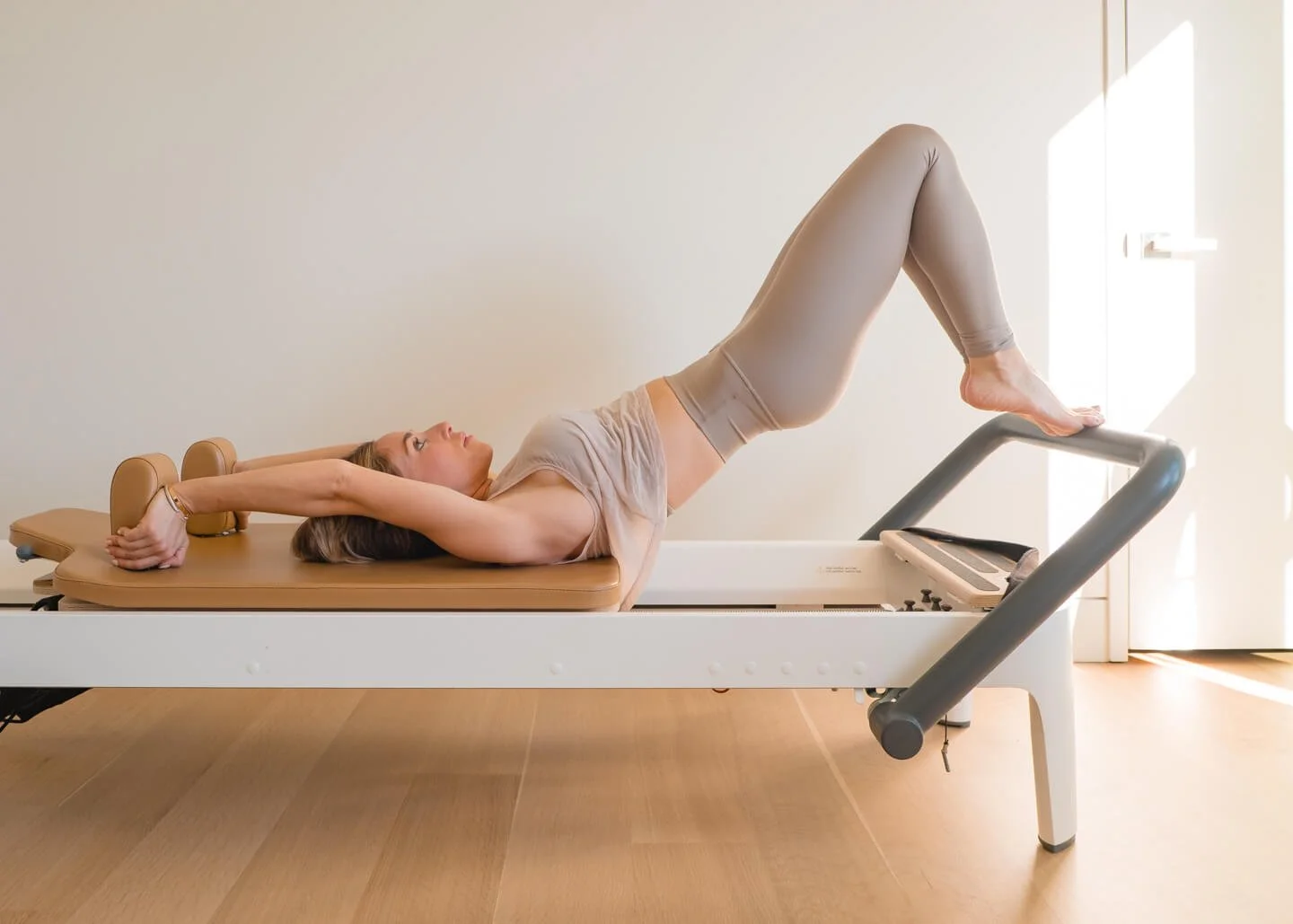 Woman lying on a Pilates reformer machine in a Pilates studio, with her legs bent and feet on the machine's footrest, performing a stretching exercise.