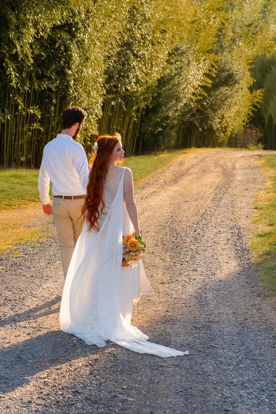 A bride and groom walking along a dirt path surrounded by green trees on a sunny day.
