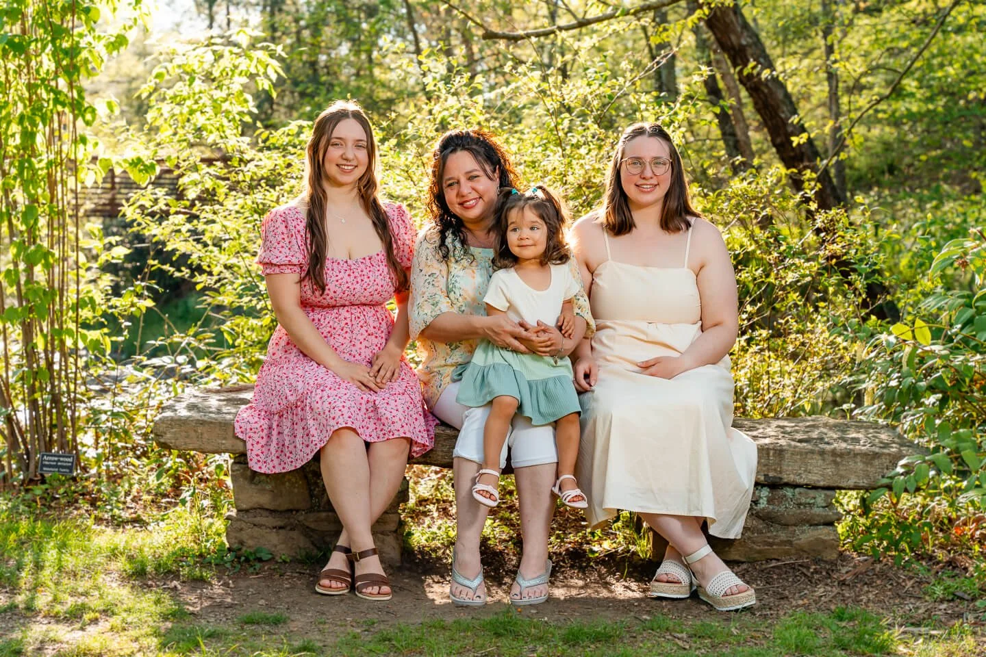 Four females sitting on a wooden bench in a forest with green foliage, smiling at the camera, dressed in light summer clothing.