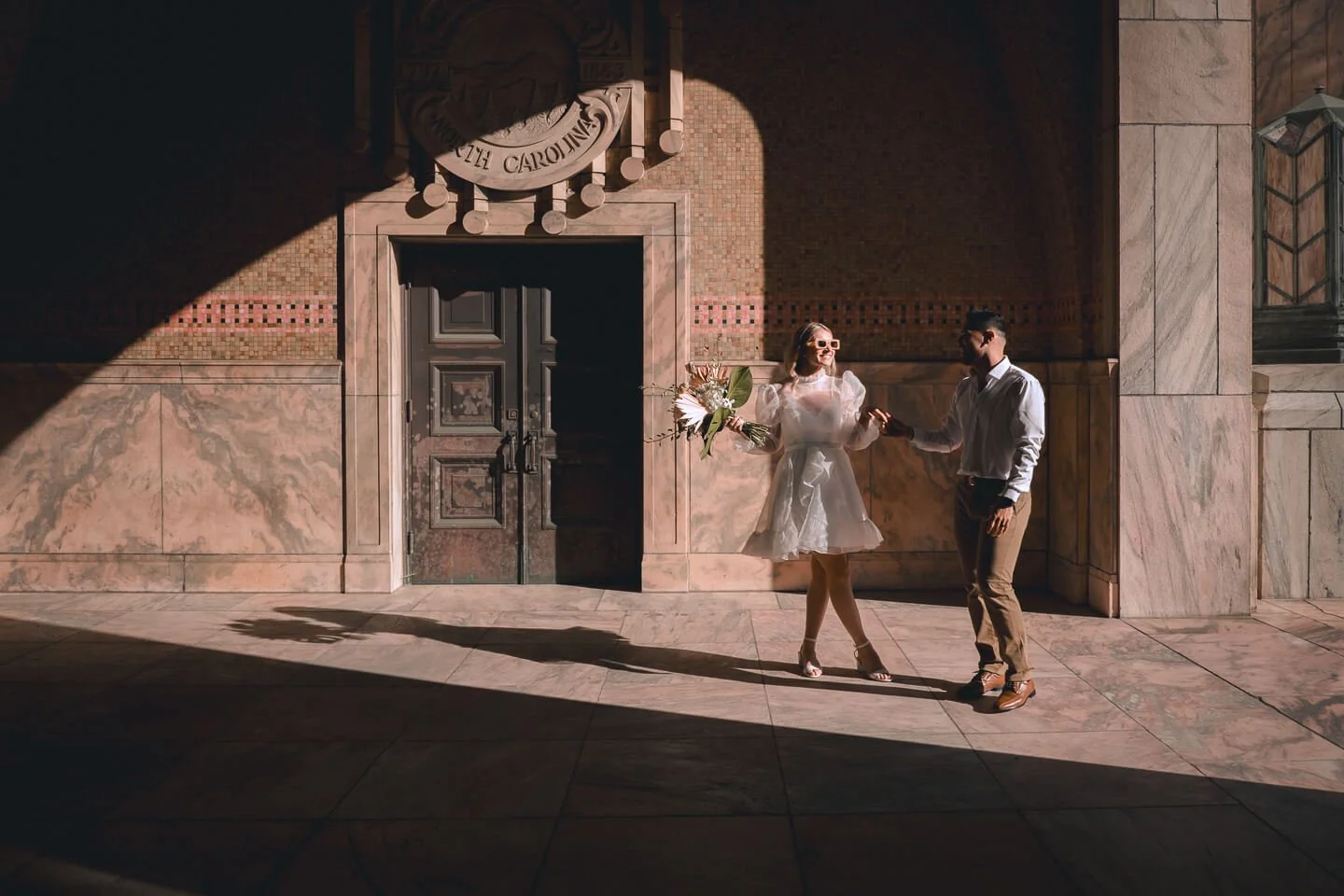 A couple dancing outdoors in front of a historic building, with dramatic sunlight creating a shadow on the wall behind them. The woman in a white dress holds a bouquet and wears sunglasses, while the man in a white shirt and brown pants holds her han