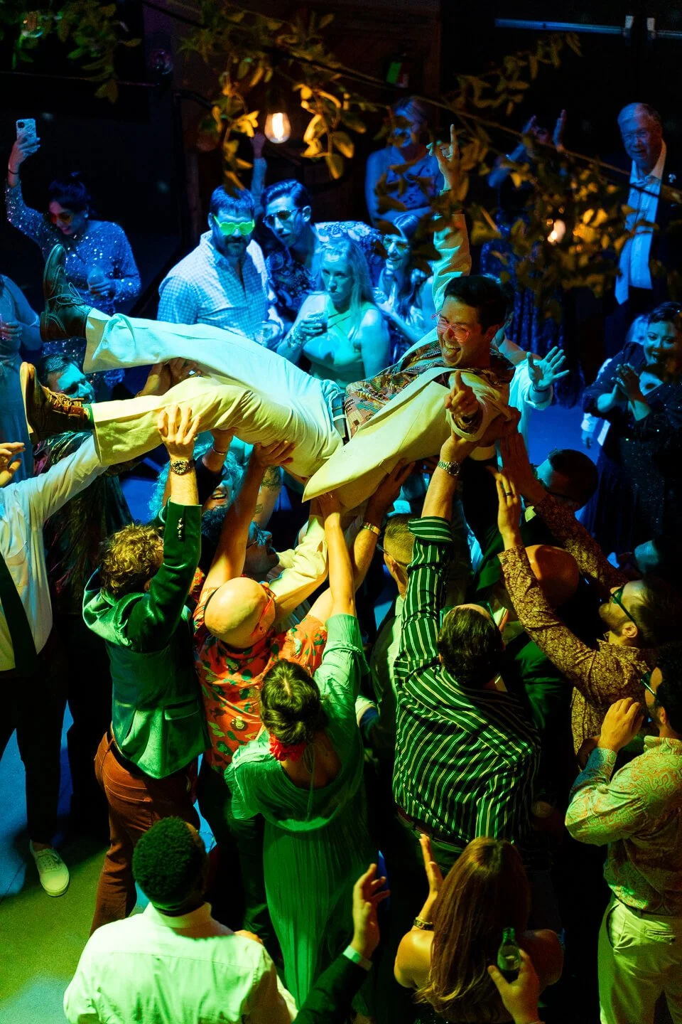 Group of people at a lively party lifting a man into the air, with colorful lighting and many guests dancing and celebrating.