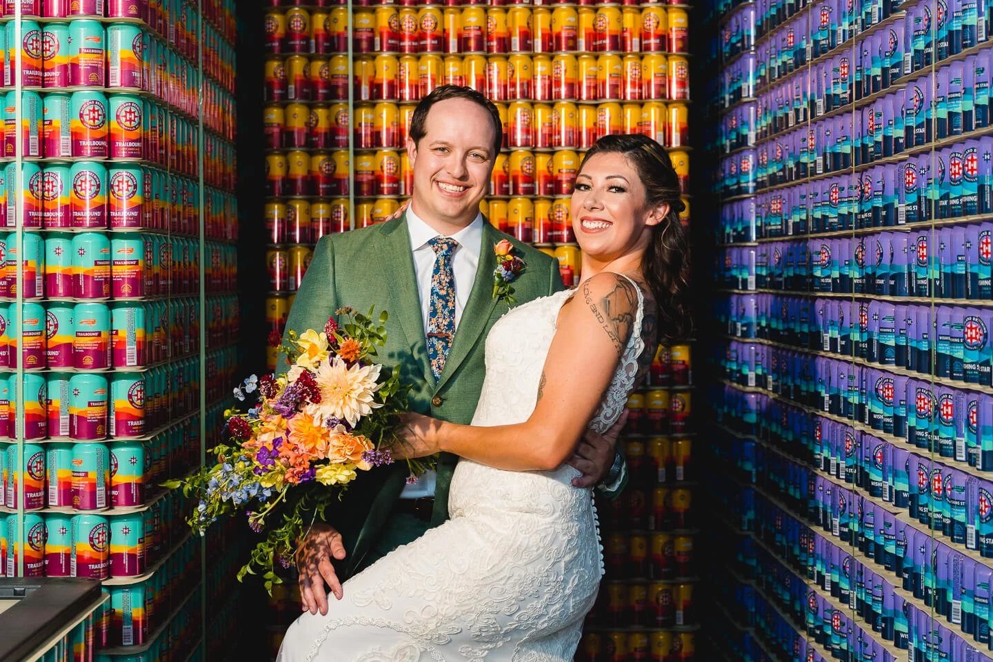 A happy couple in wedding attire standing in a room with shelves filled with colorful cans. The groom is wearing a green suit and floral tie, holding a bouquet of flowers. The bride is dressed in a white lace wedding gown with tattoos visible on her