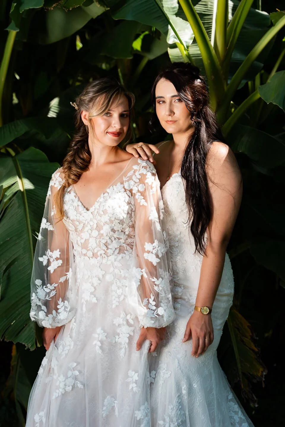 Two women dressed in wedding gowns standing in front of lush green foliage.