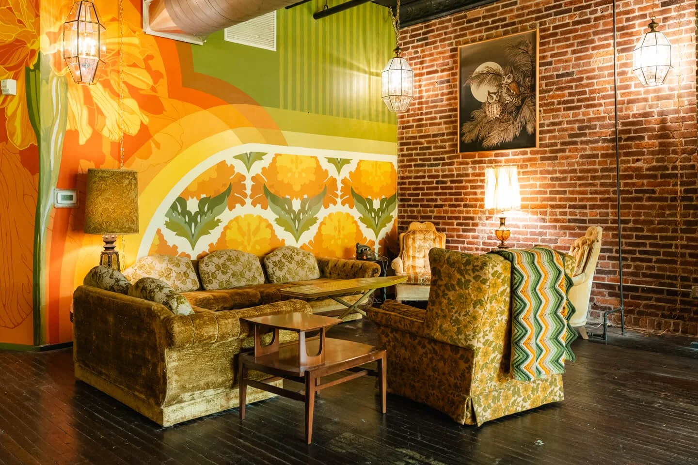 Colorful retro living room with floral painted wall, vintage furniture, a brick wall, and hanging pendant lamps.