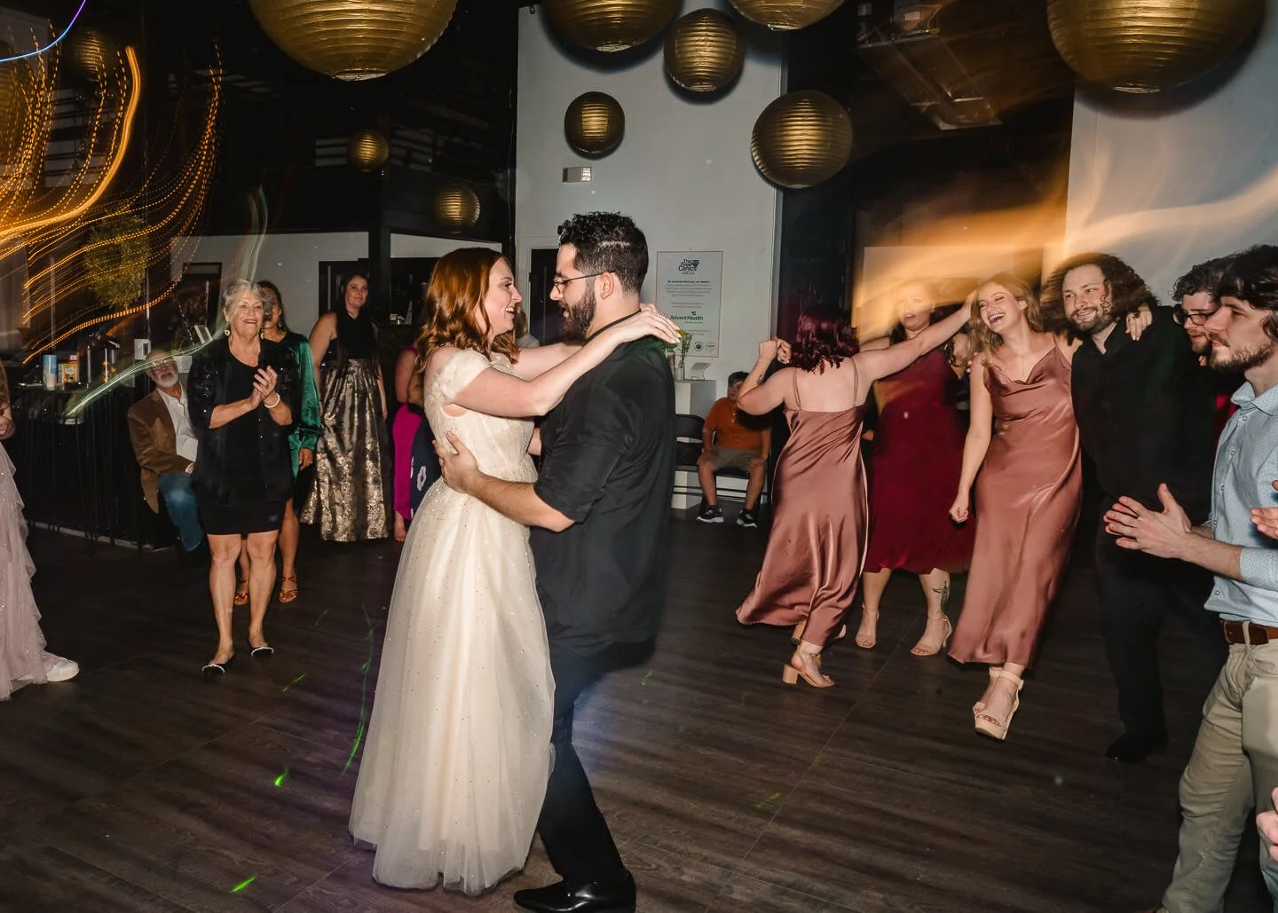 Couple dancing at a wedding reception with guests in the background, some clapping and smiling, illuminated by hanging paper lanterns and colorful lights.