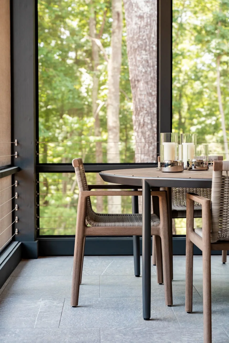Round dining table with three candles in glass holders and modern chairs in front of a large window showing a view of green trees outside.