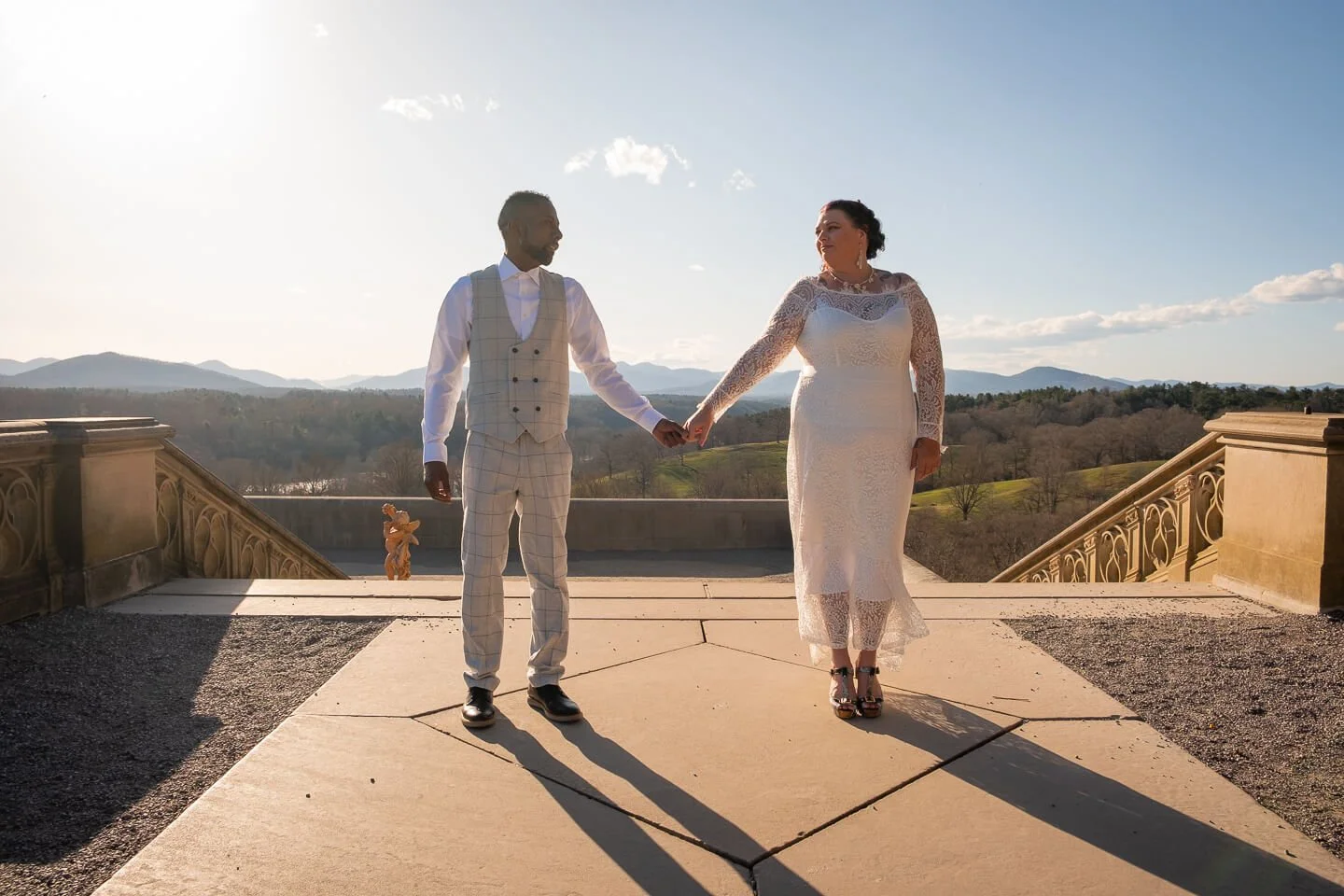 A bride and groom holding hands outdoors on a stone terrace with scenic landscape and mountains in the background