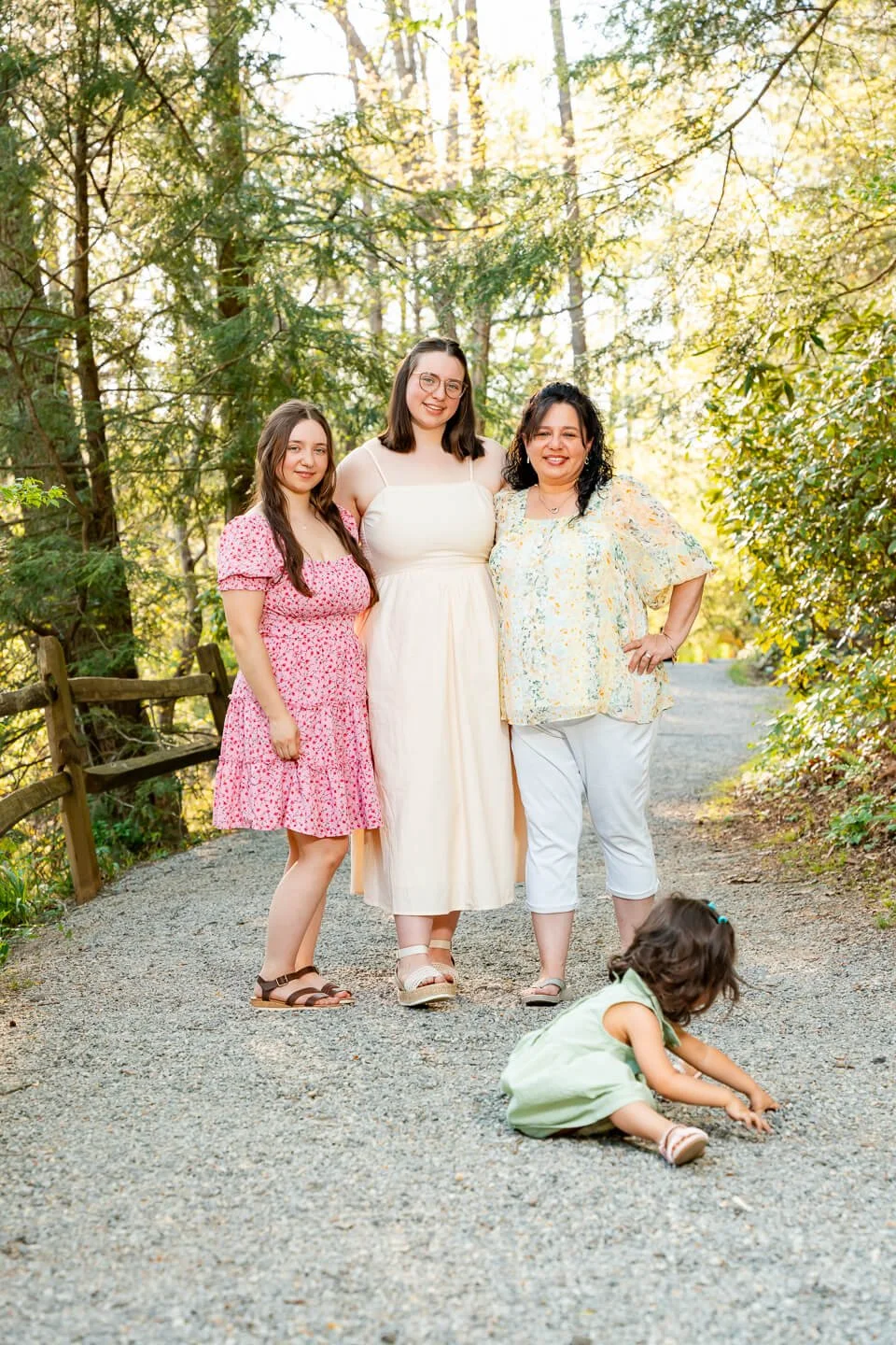 Three women and a young girl standing on a gravel path in a wooded area during daytime, smiling at the camera, with another young girl sitting on the ground in front of them.
