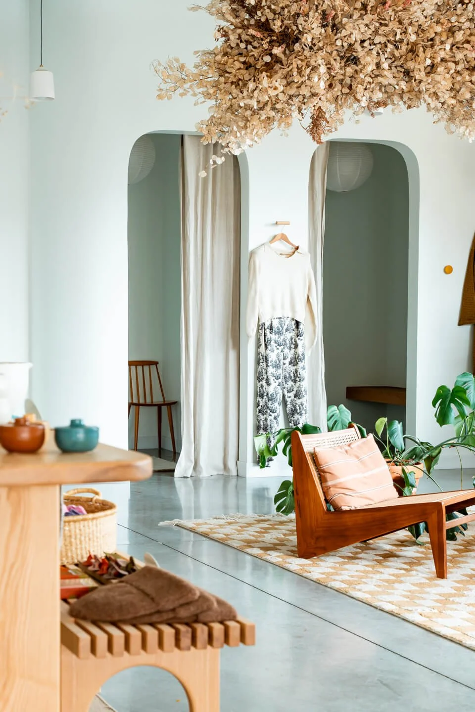 A cozy, modern living space with a display of clothing hanging on a wall, a wooden bench with cushions, potted plants, and soft lighting.