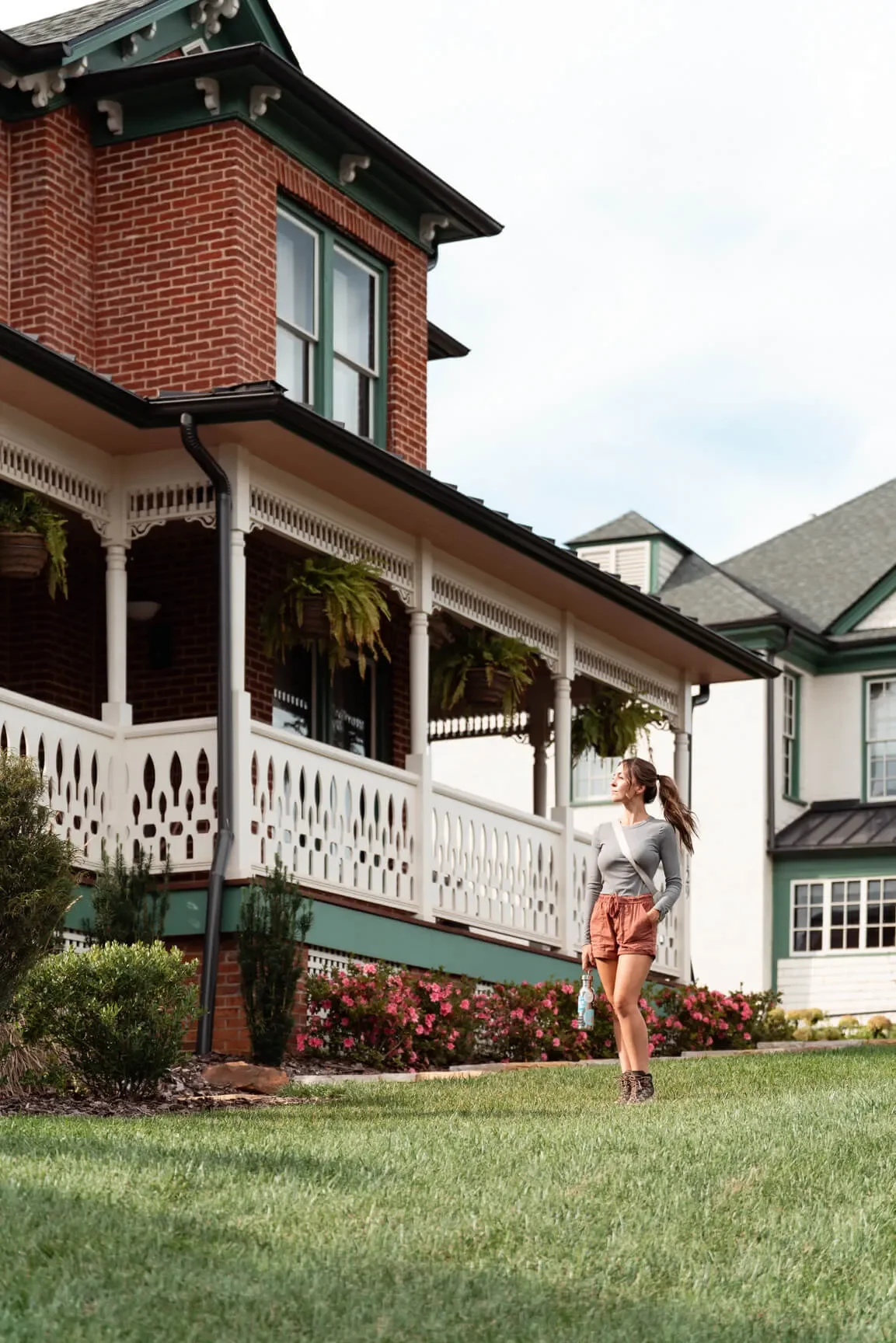 A young woman with brown hair tied in a ponytail wearing a light gray long-sleeve shirt, orange shorts, and hiking boots, standing on a well-kept lawn next to a large historic house with a white porch, red brick walls, and hanging plants.