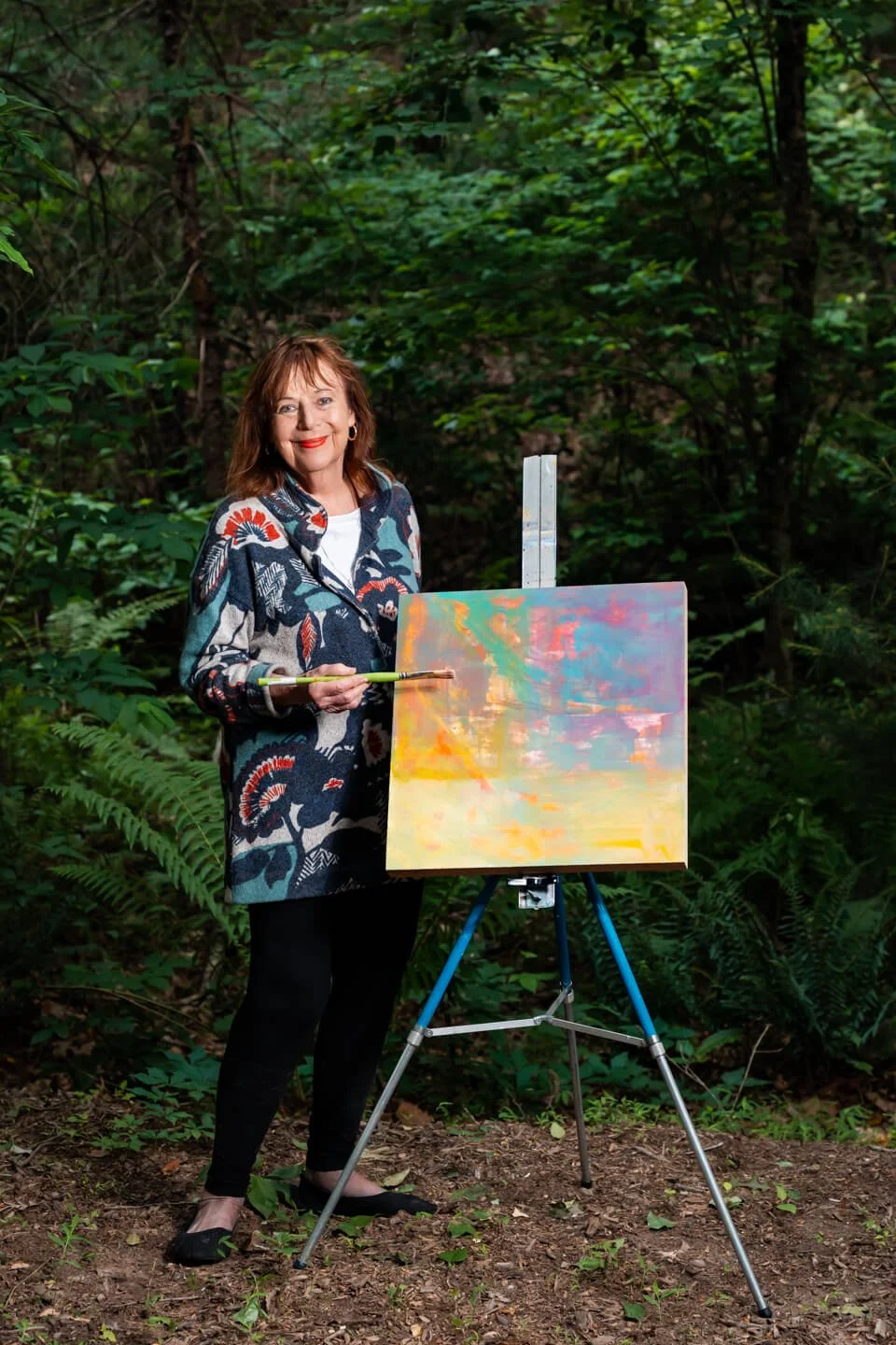 A woman with shoulder-length auburn hair wearing a patterned coat and black pants stands outdoors in a wooded area, holding a paintbrush in her hand, next to an easel with a colorful abstract painting.