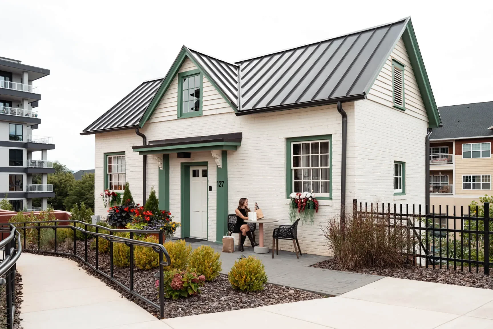 A woman sitting outside a white brick house with green trim, flowers, and a small porch, on a cloudy day.