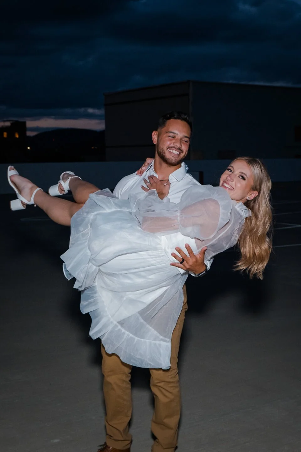 A man in white shirt and tan pants carries a smiling woman in a white dress and heels in a parking lot at night against a dark, cloudy sky.