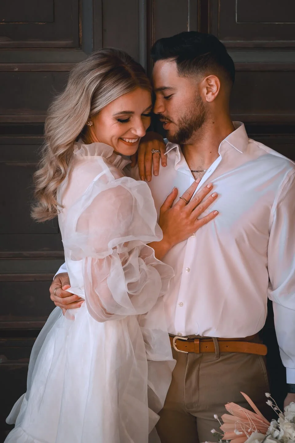 A happy couple in wedding attire sharing an intimate moment, with their faces close and eyes closed, smiling softly against a dark wooden background.