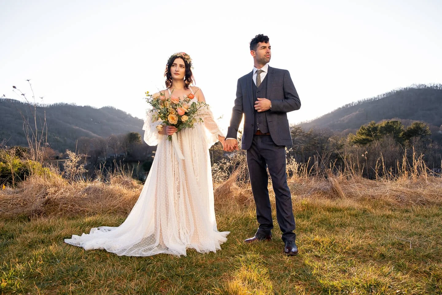 A bride and groom holding hands outdoors on a grassy field with mountains in the background during sunset.