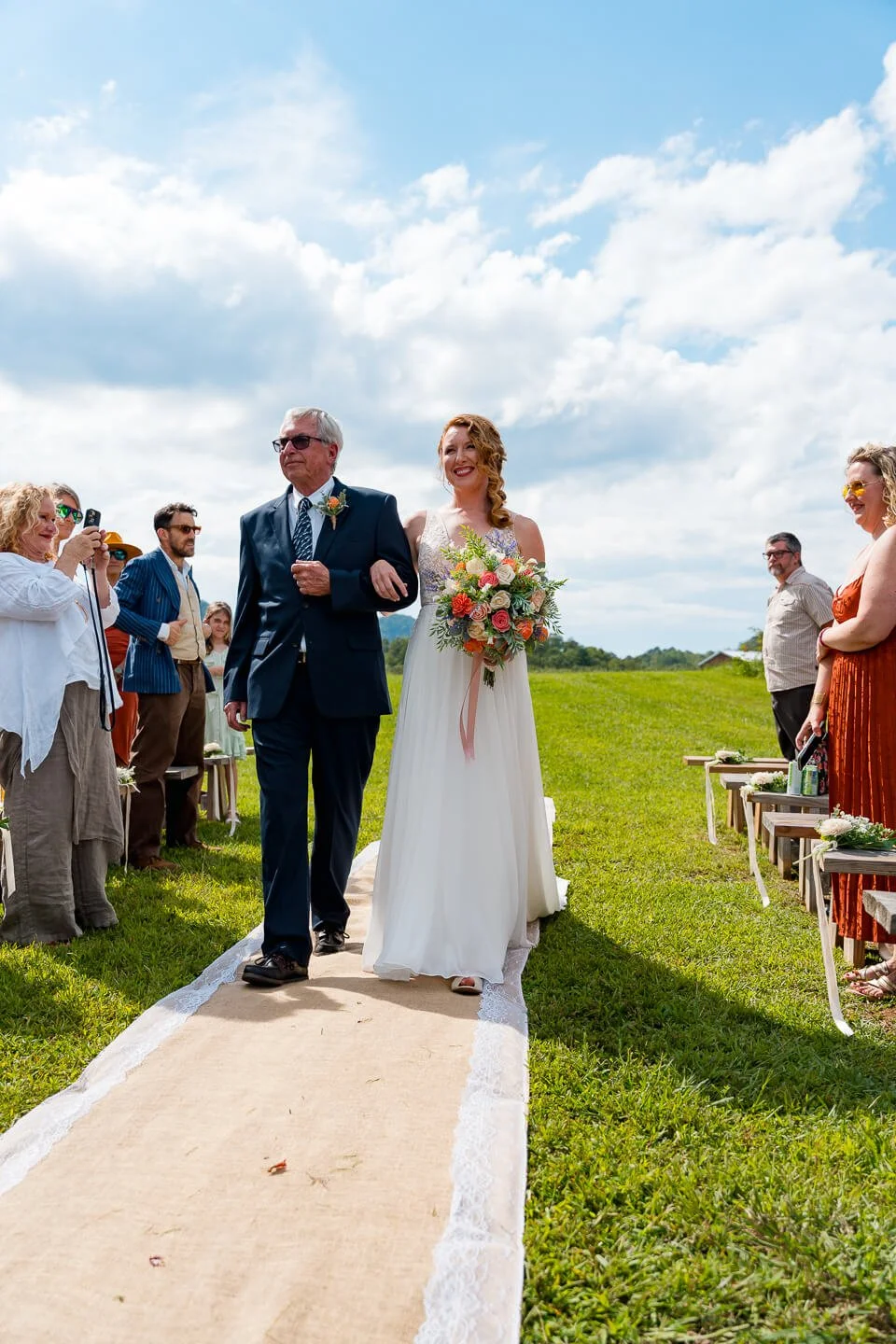 A bride walking down the aisle on an outdoor wedding ceremony with her father, holding a bouquet, surrounded by guests on a grassy field under a partly cloudy sky.