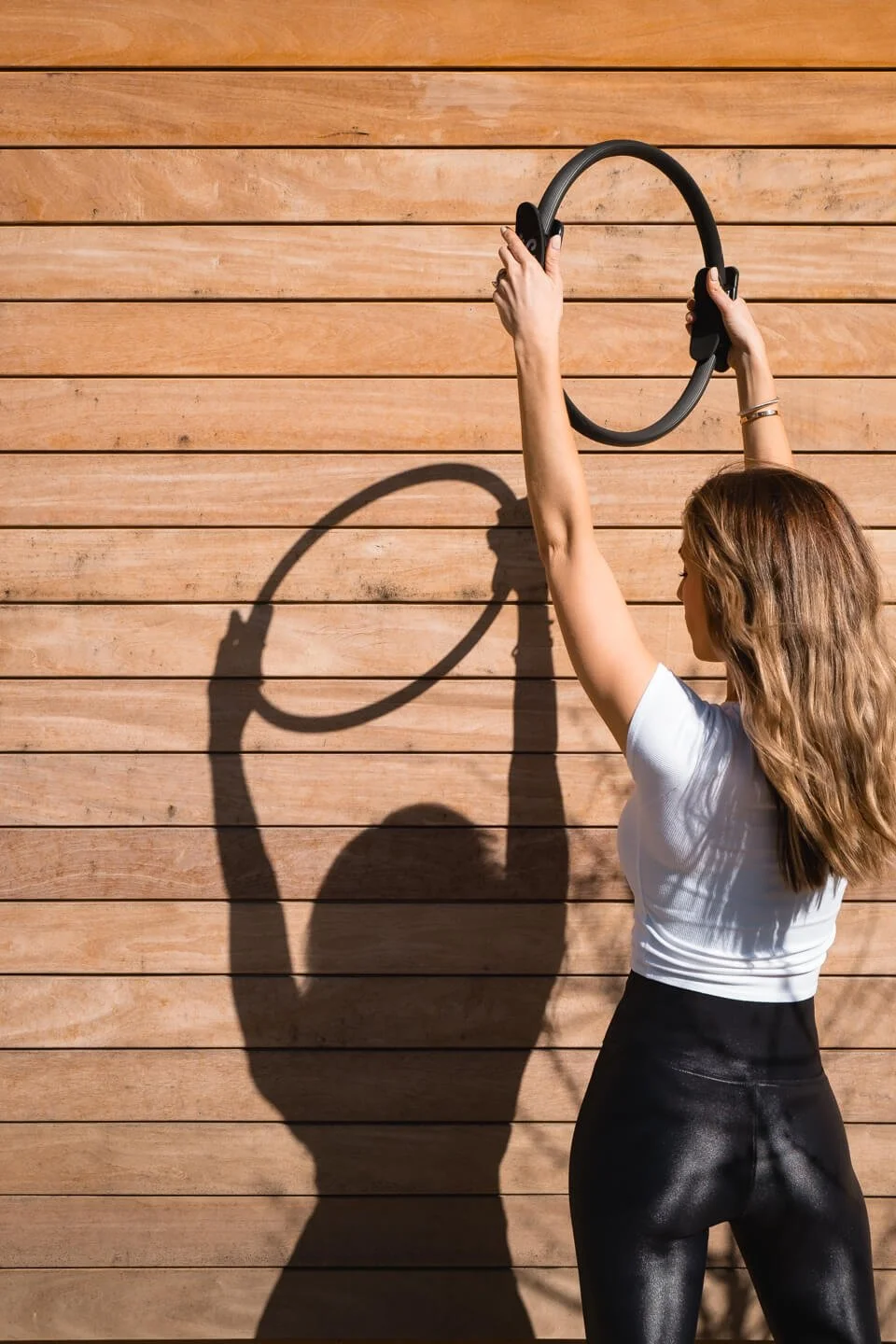 A woman with wavy light brown hair, wearing a white top and black shiny pants, is holding a circular exercise ring above her head against a wooden-paneled wall. Her shadow is cast on the wall.