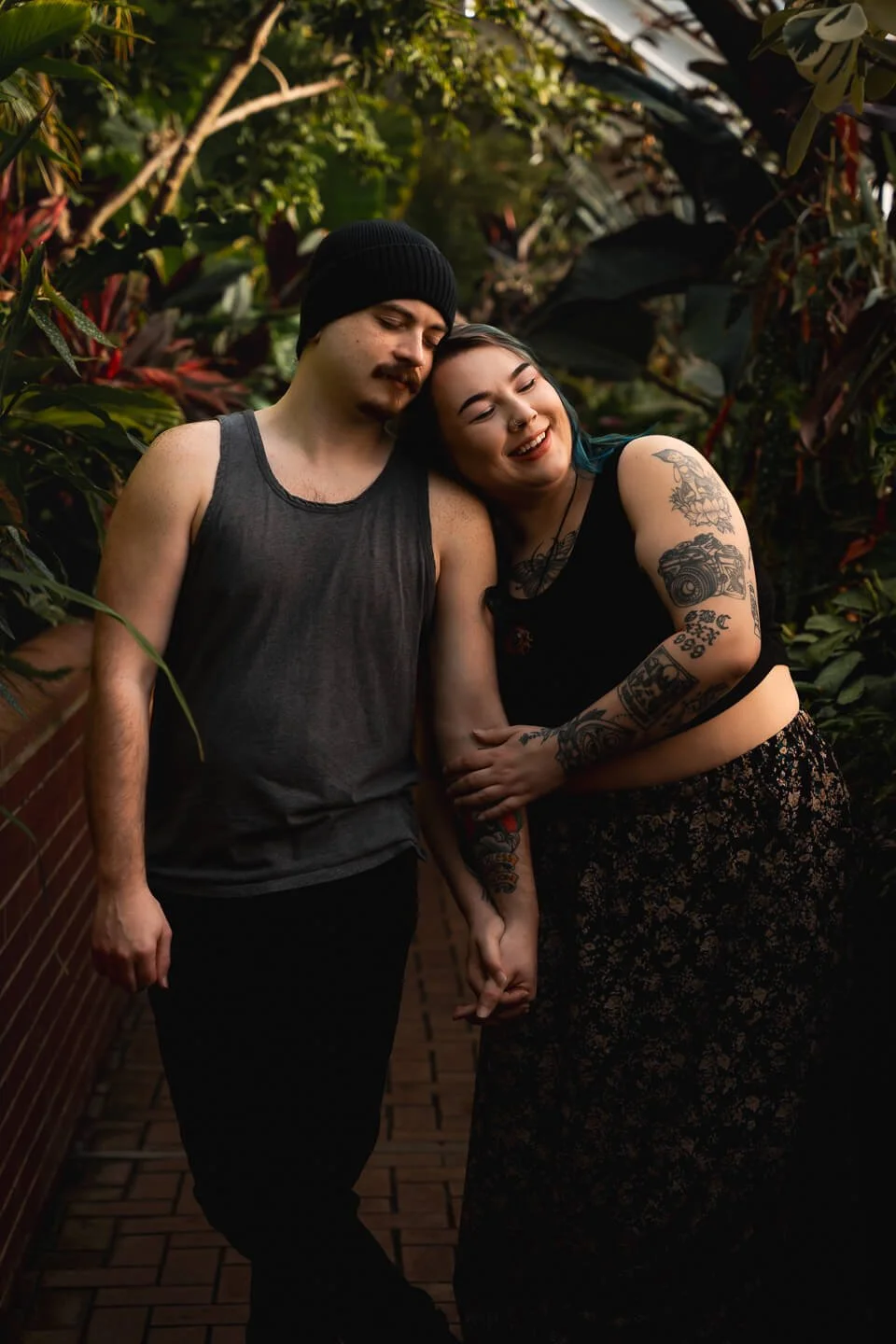 A young couple stands close together in a lush, green botanical garden. The woman has tattoos on her arms and is smiling with a joyful expression, resting her head on the man's shoulder. The man wears a black beanie and a gray tank top, with a serene