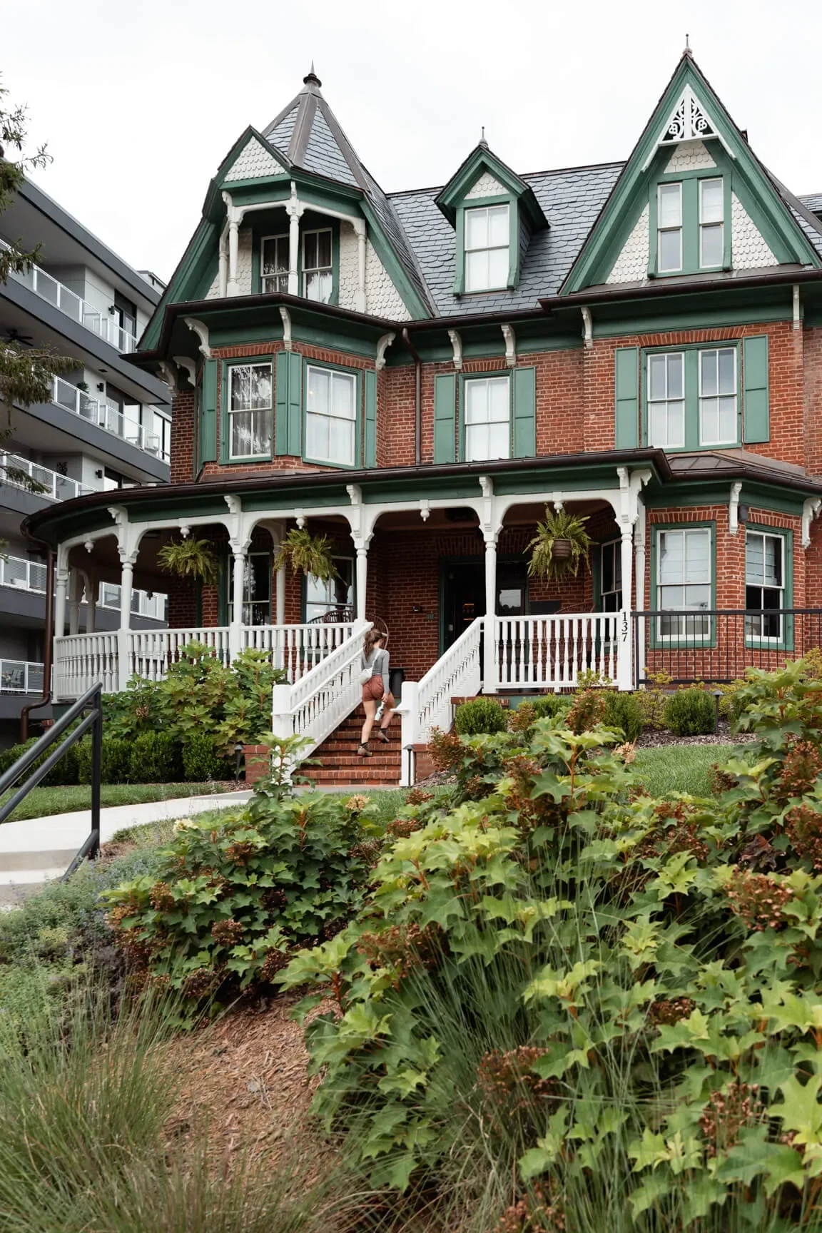 A large Victorian-style house with red brick walls, green window shutters, and a steeply pitched roof. The house has a white front porch with stairs and hanging plants. A person is climbing the stairs to the entrance, and there are lush green shrubs