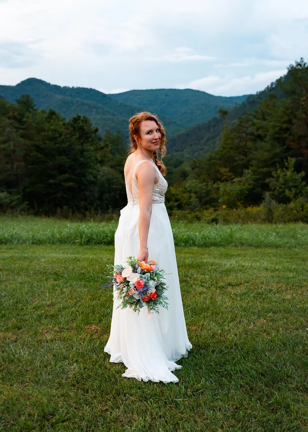 A woman in a white wedding dress standing on grassy field with mountains and trees in the background, holding a bouquet of pink, white, and orange flowers, looking back over her shoulder.