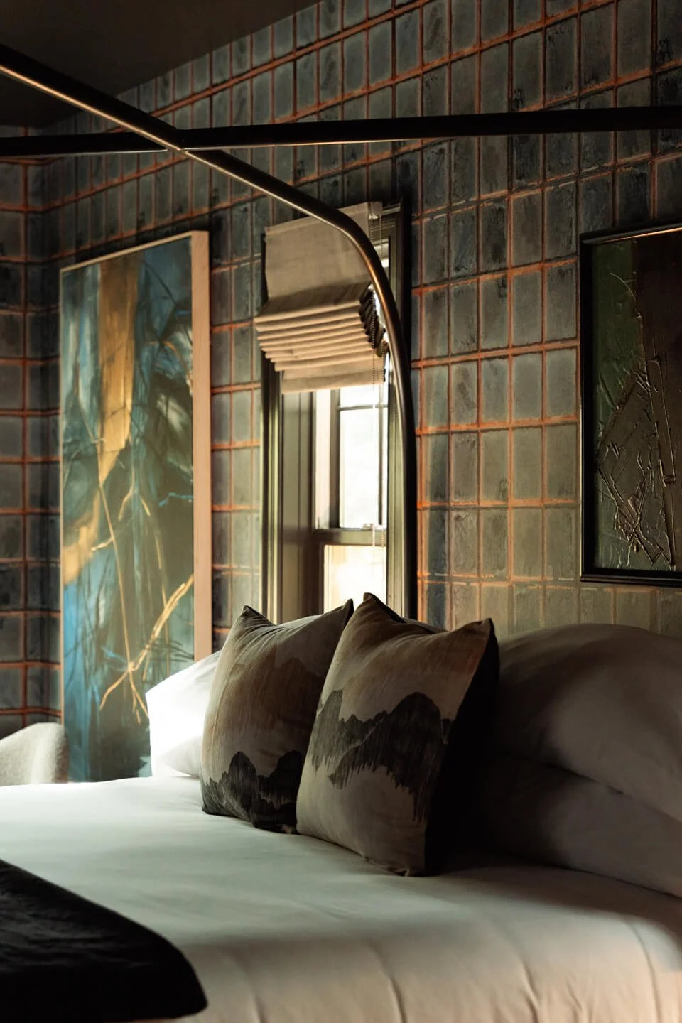 A cozy bedroom with a bed featuring two decorative pillows with mountain landscape prints, set against a wall with industrial-style brick tiles, a window with a Roman shade, and abstract artwork on the walls.