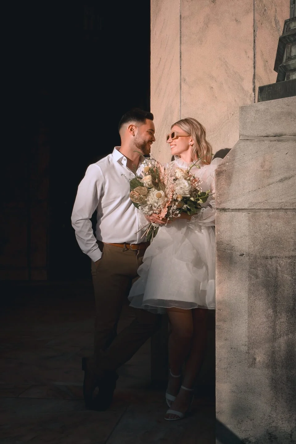 A smiling bride and groom in wedding attire sharing a moment together. She holds a bouquet of flowers, and he has his hand in his pocket, leaning against a stone wall.