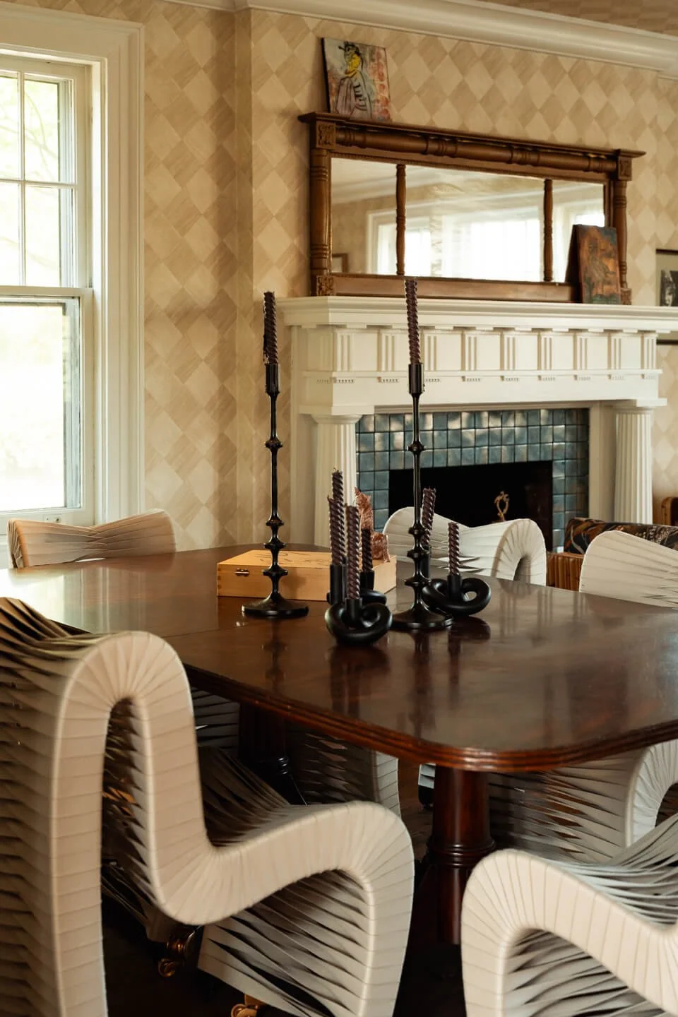 Dining room with a dark wood table, white modern chairs, black candlesticks, and a fireplace with a mirror and paintings above it.