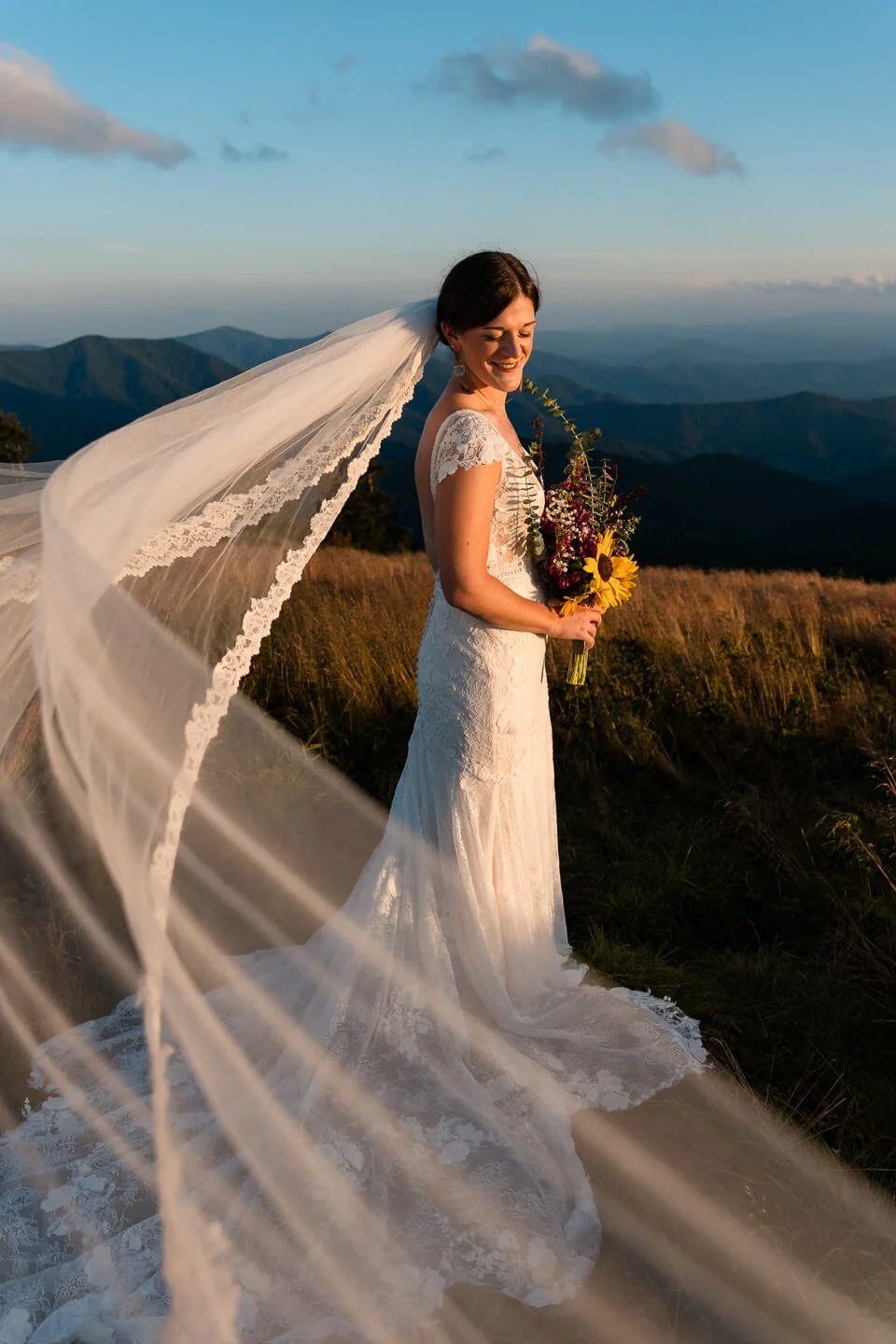 A bride in a lace wedding dress holding a bouquet of flowers, standing outdoors on a mountain during sunset, with her veil flowing in the wind and scenic mountains in the background.