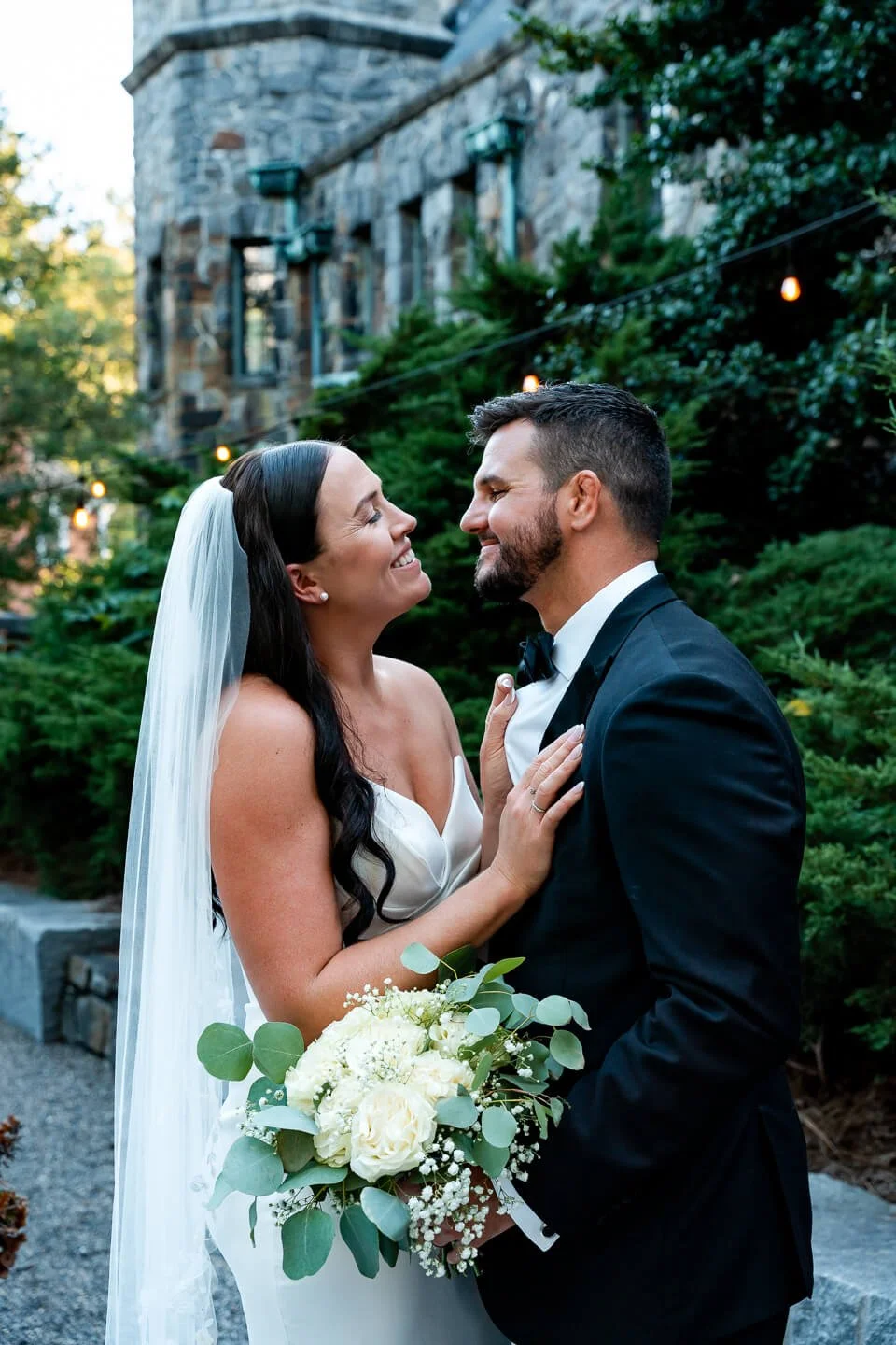 Bride and groom smiling and looking at each other outdoors, woman in a white wedding dress holding a bouquet of white roses and greenery, man in a black tuxedo, surrounded by greenery and a stone building in the background.