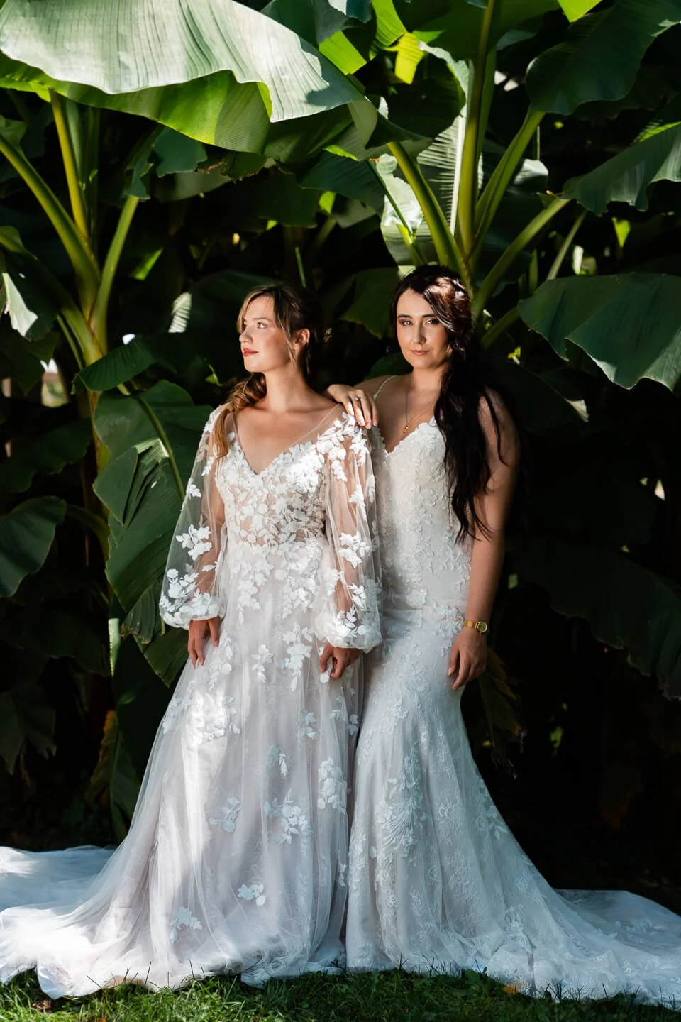 Two women in wedding dresses standing outside in front of large green leaves.