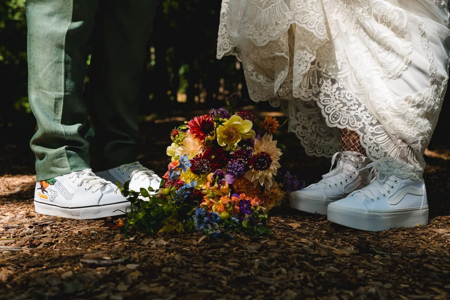 Close-up of a wedding bouquet laying on the ground between two people wearing sneakers, one in a lace dress and the other in suit pants, with a forest background.