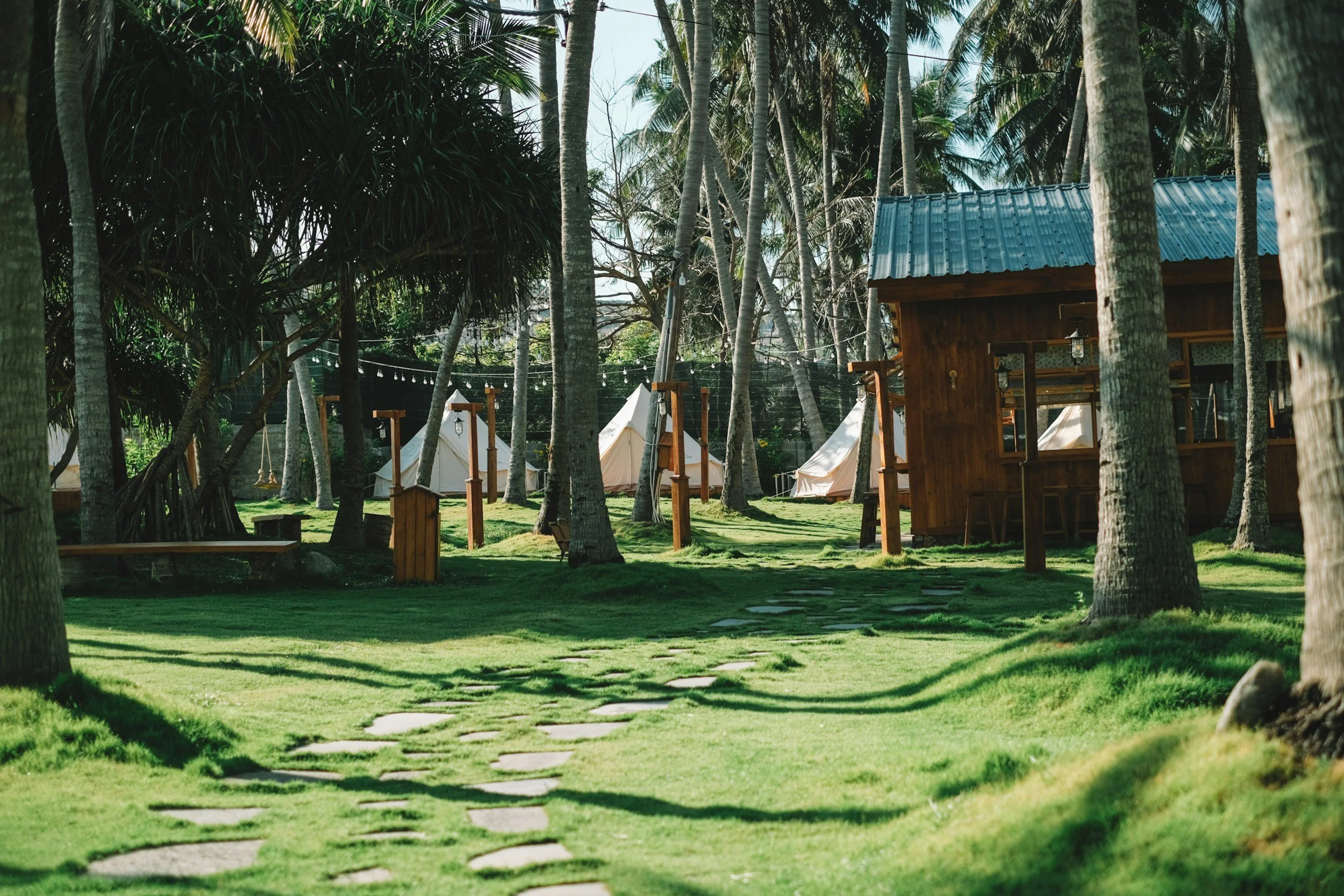 A tropical outdoor scene with wooden cabins, white tents, and tall palm trees. A stone pathway winds through a grassy area with structures and hanging swings.