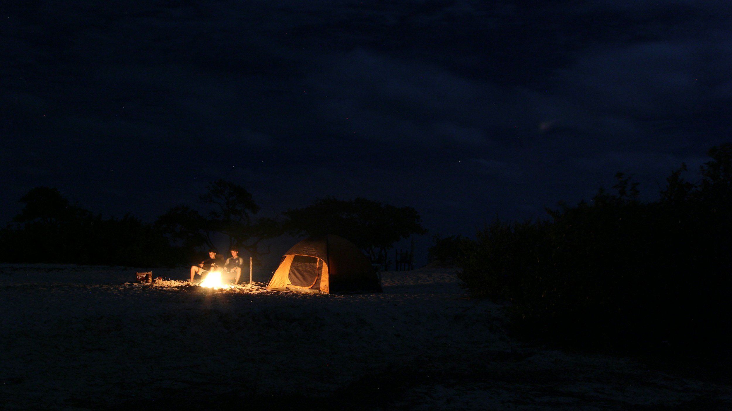 Two people sitting by a campfire near a tent on a sandy landscape at night, with trees in the background and a starry sky above.